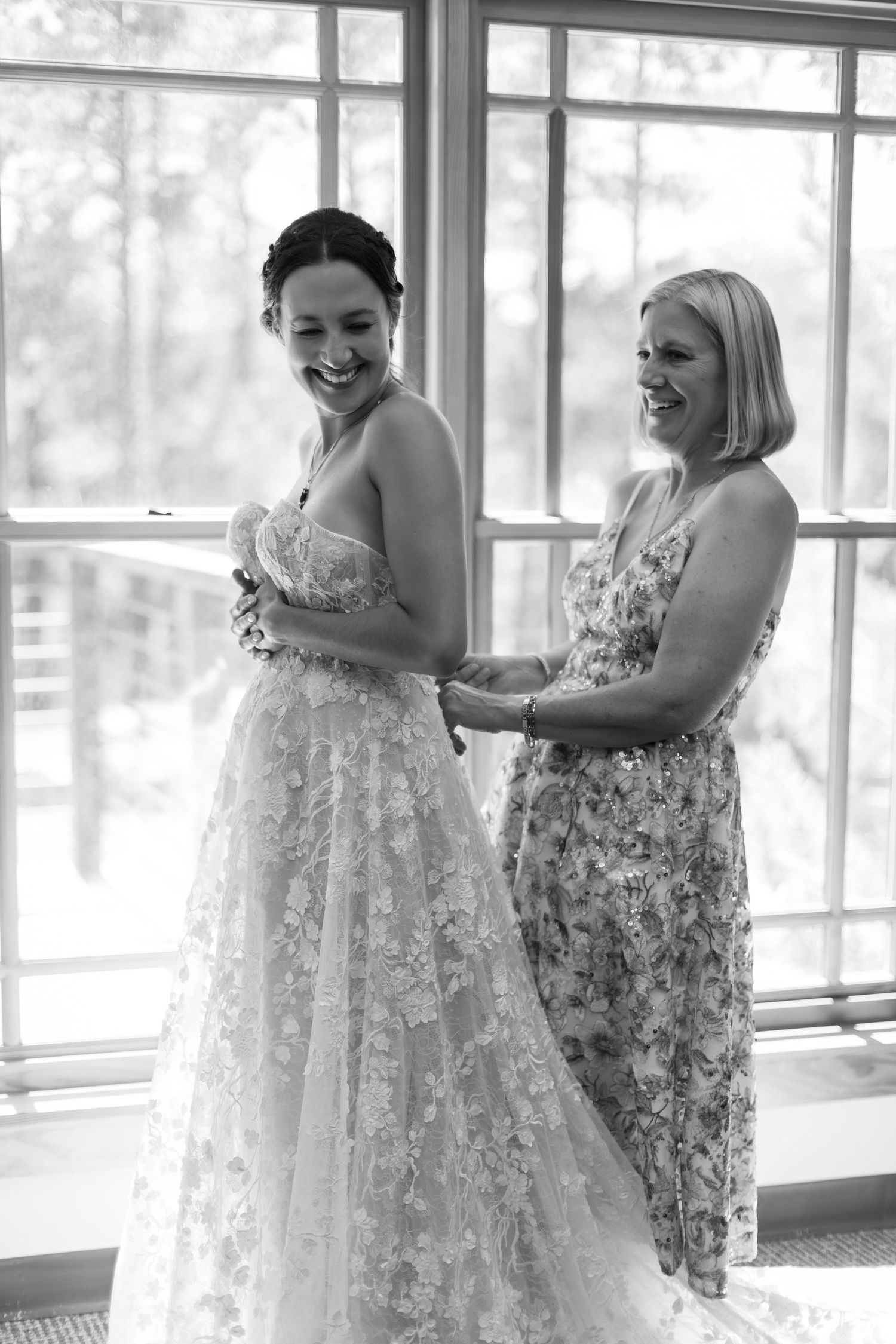 Bride laughing in lace wedding gown while woman helps her dress, black and white photo near large windows.