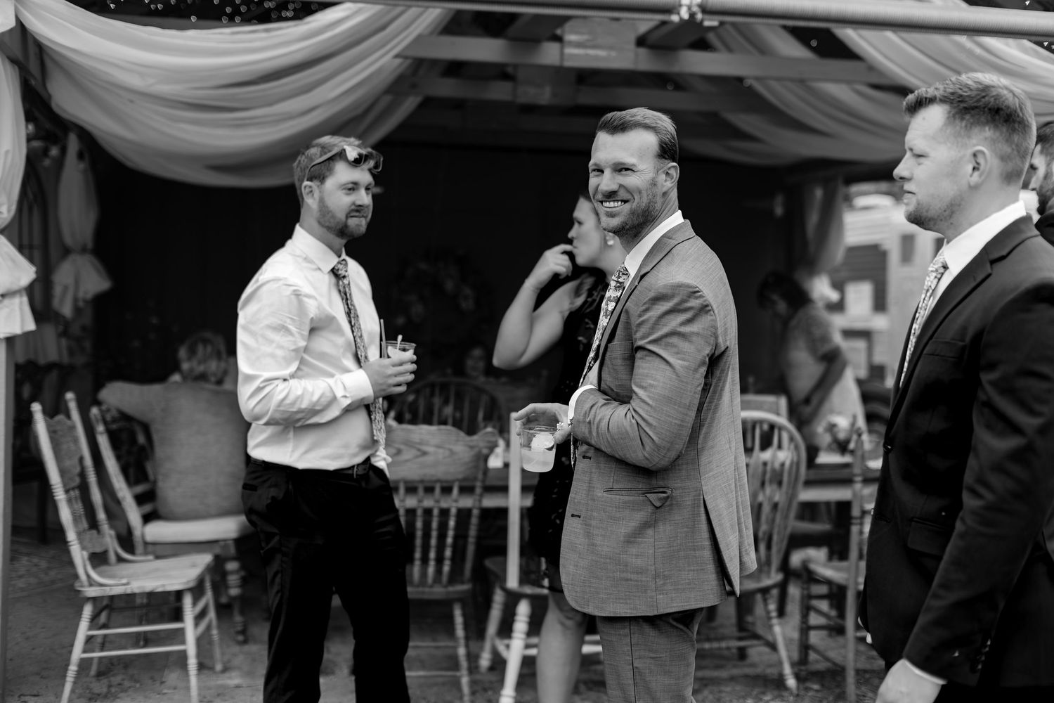 Three men in formal attire conversing and laughing at an outdoor wedding reception in black and white photo.
