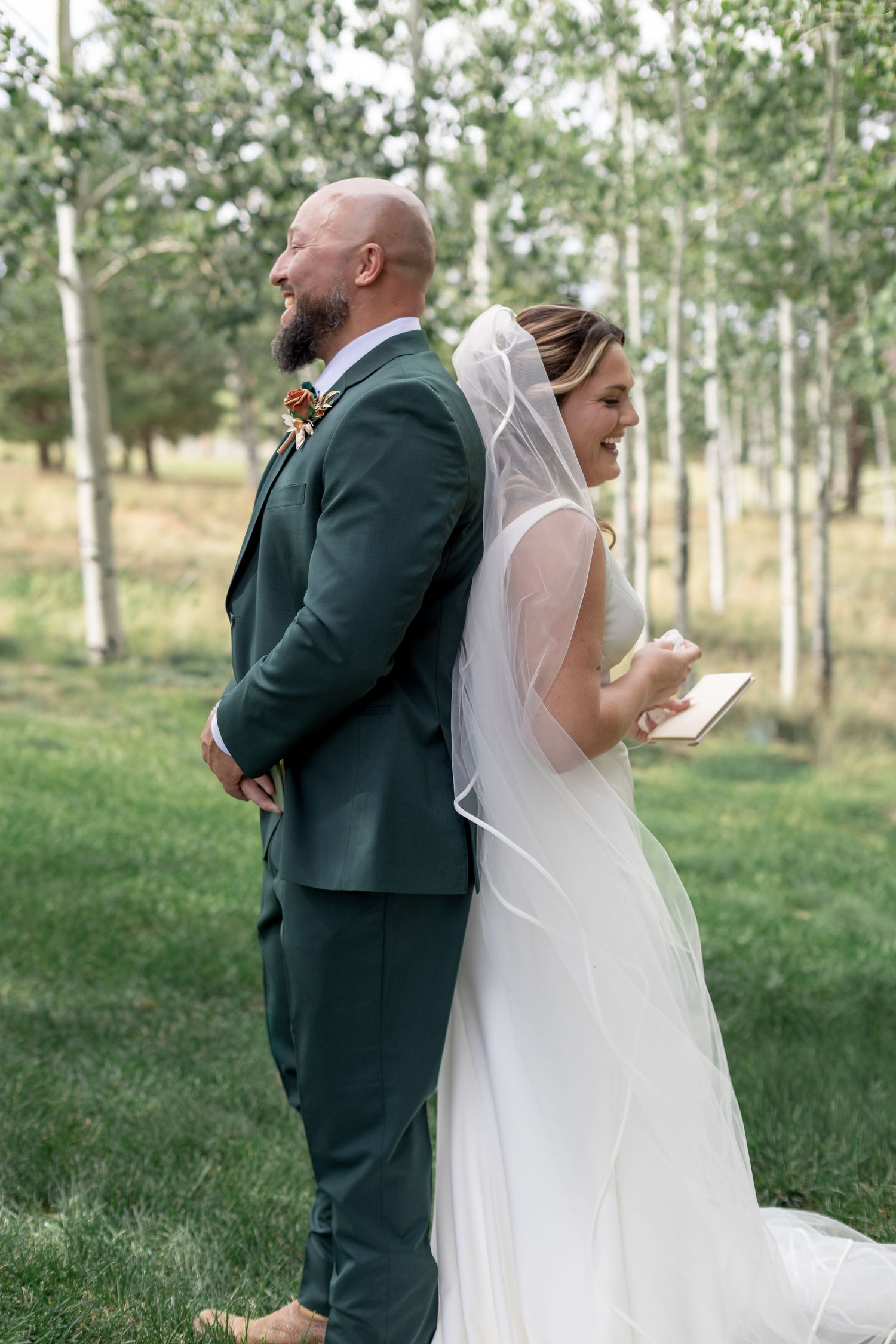 Bride and groom stand back-to-back in forest reading vows, bride in white gown with veil, groom in dark green suit.
