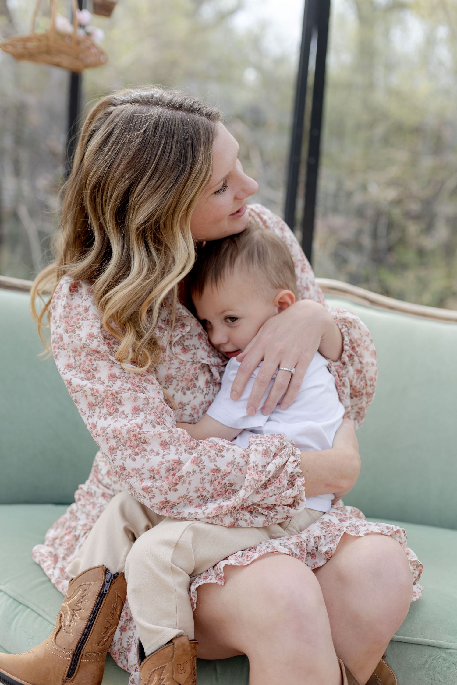 A mother in a floral dress hugs her toddler on a mint green sofa, sharing a tender embrace.