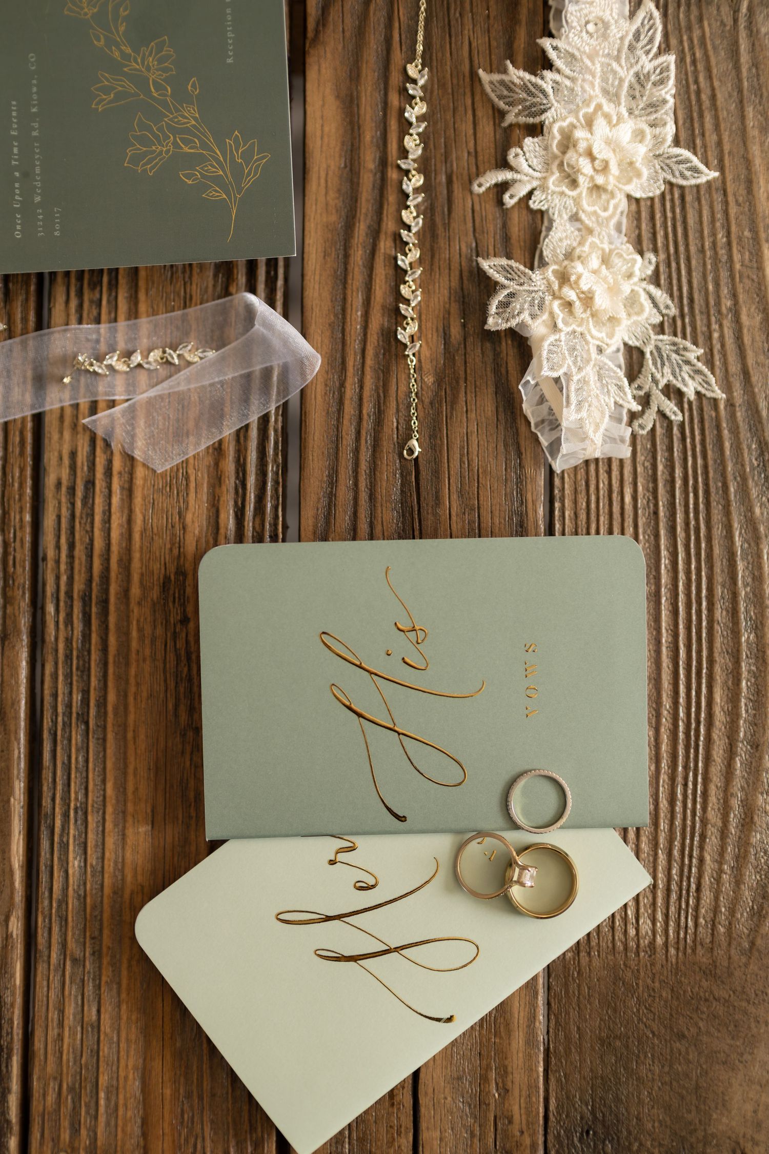 Elegant wedding flat lay with sage green His and Hers cards, gold rings, lace garter, and floral jewelry on wood.