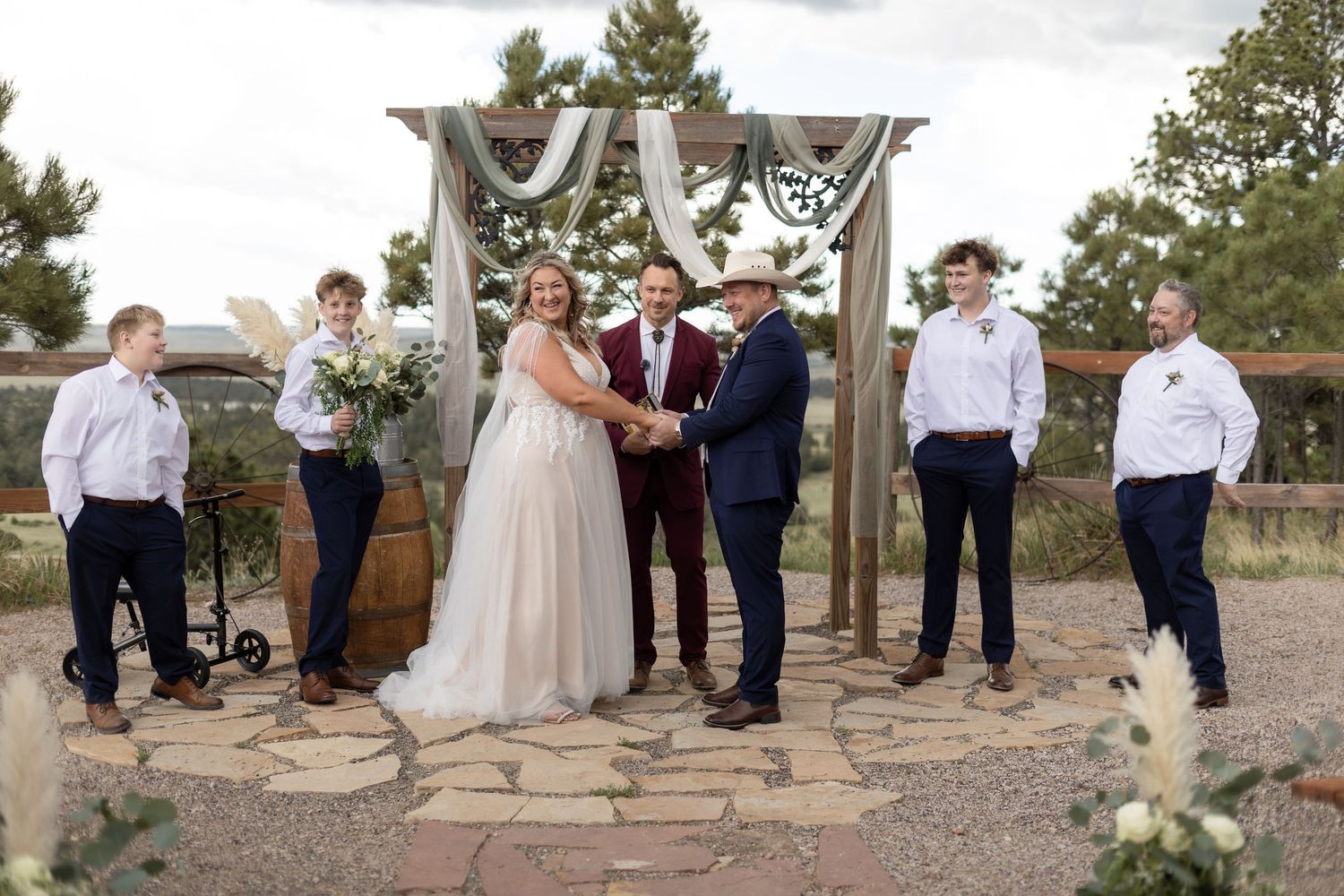 Outdoor wedding ceremony with bride and groom holding hands at rustic wooden arch, flanked by groomsmen in navy suits.