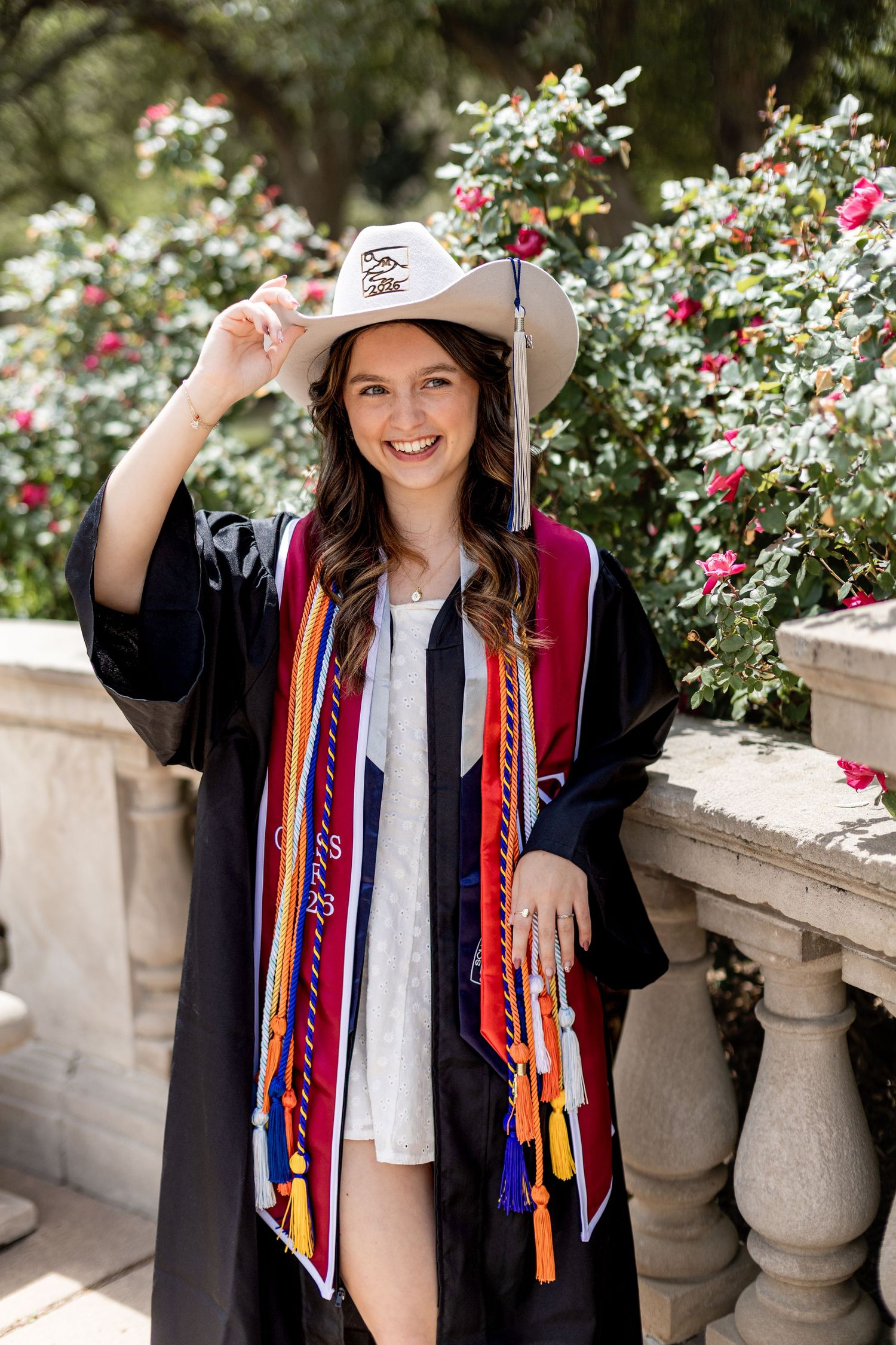 Smiling female graduate in black gown, colorful stoles, and cowboy hat posing outdoors near roses.