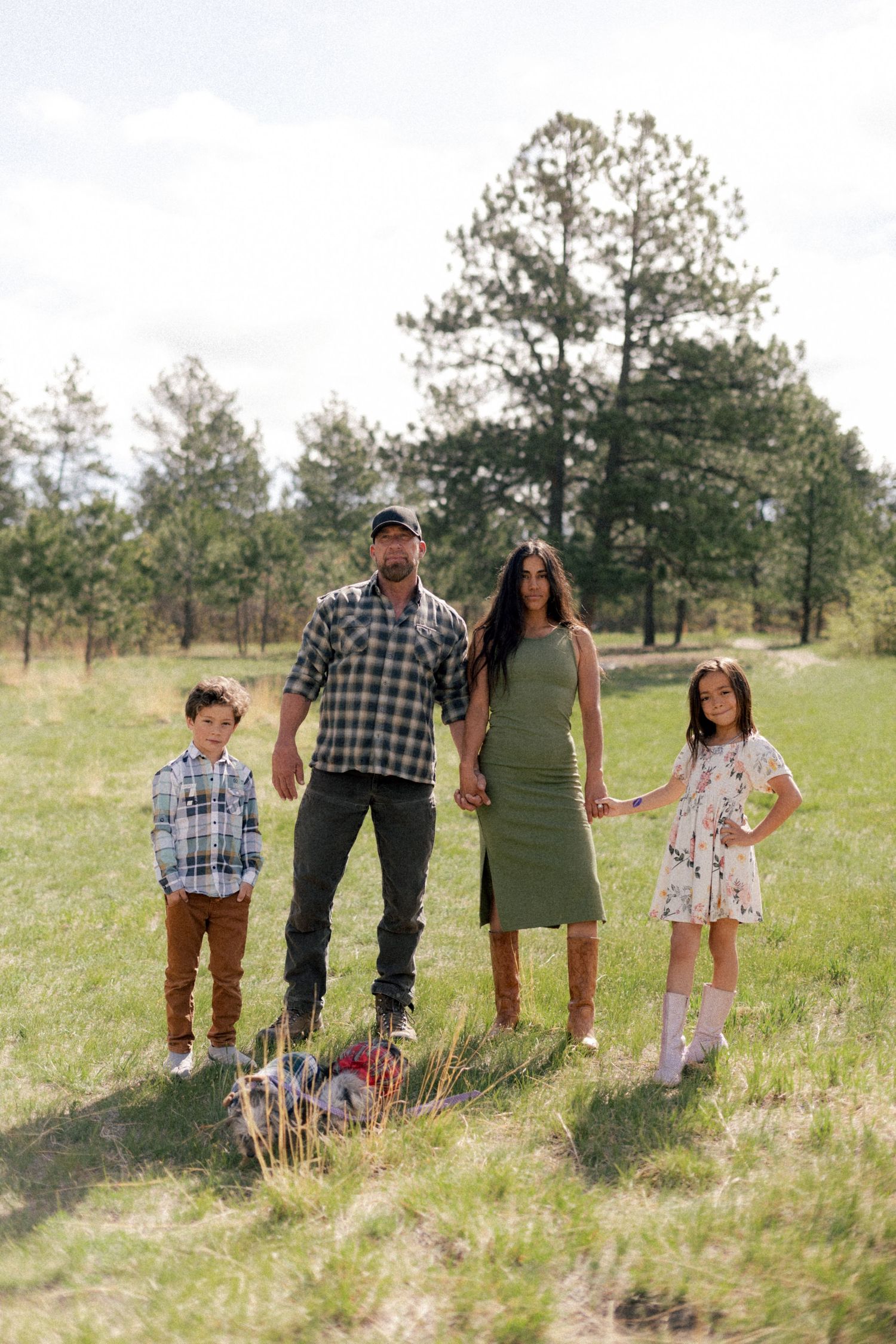 Family of four posing outdoors in a grassy field surrounded by pine trees, wearing casual country-style outfits.