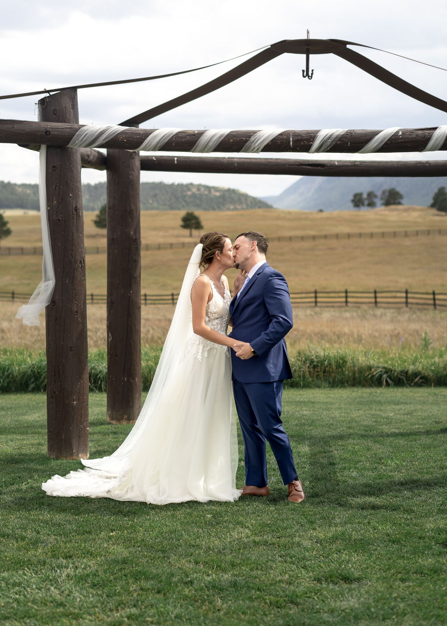 Bride and groom share a kiss under a rustic wooden arch on a green lawn with scenic mountain fields behind them.