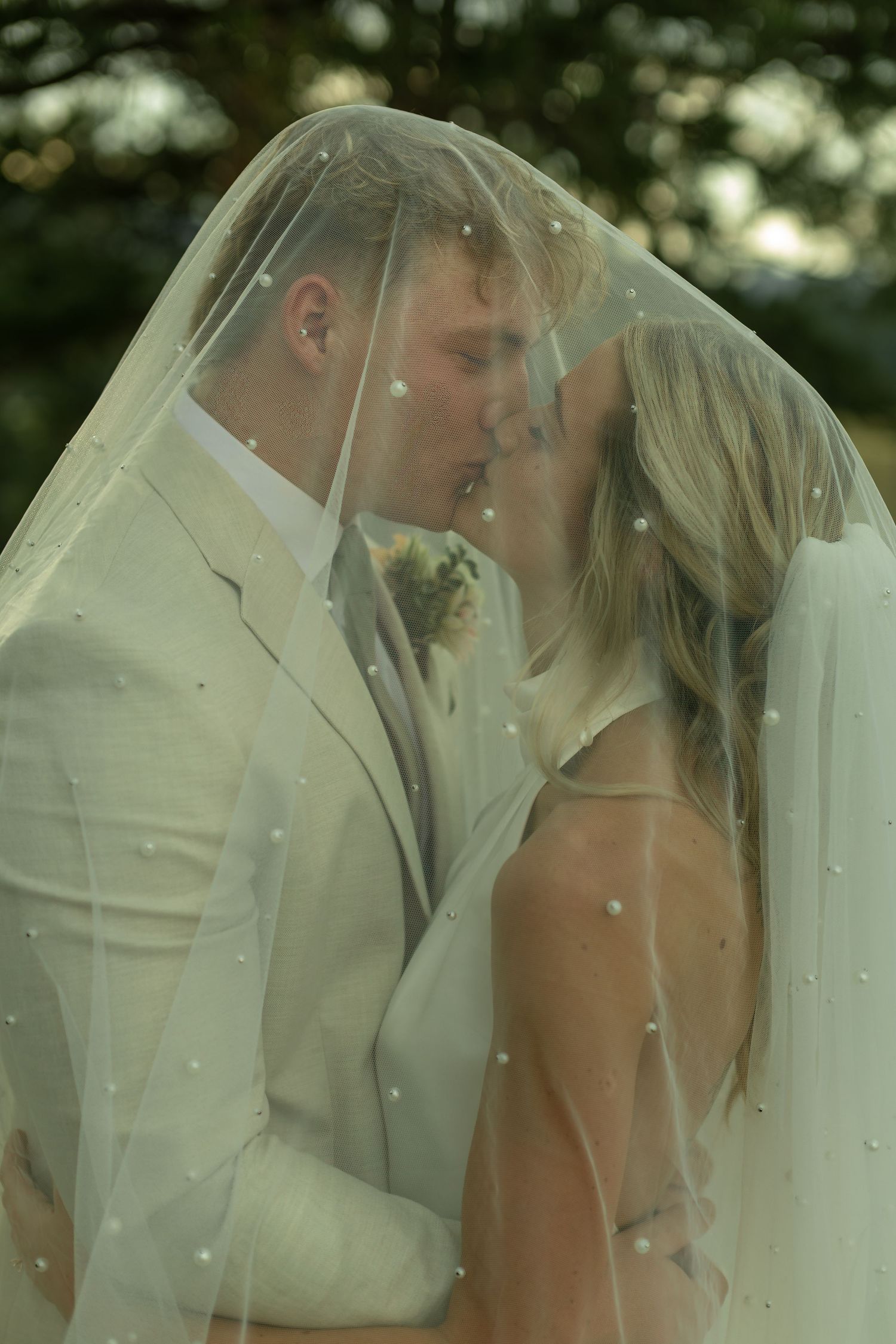 A bride and groom share a romantic kiss under a pearl-adorned wedding veil outdoors.