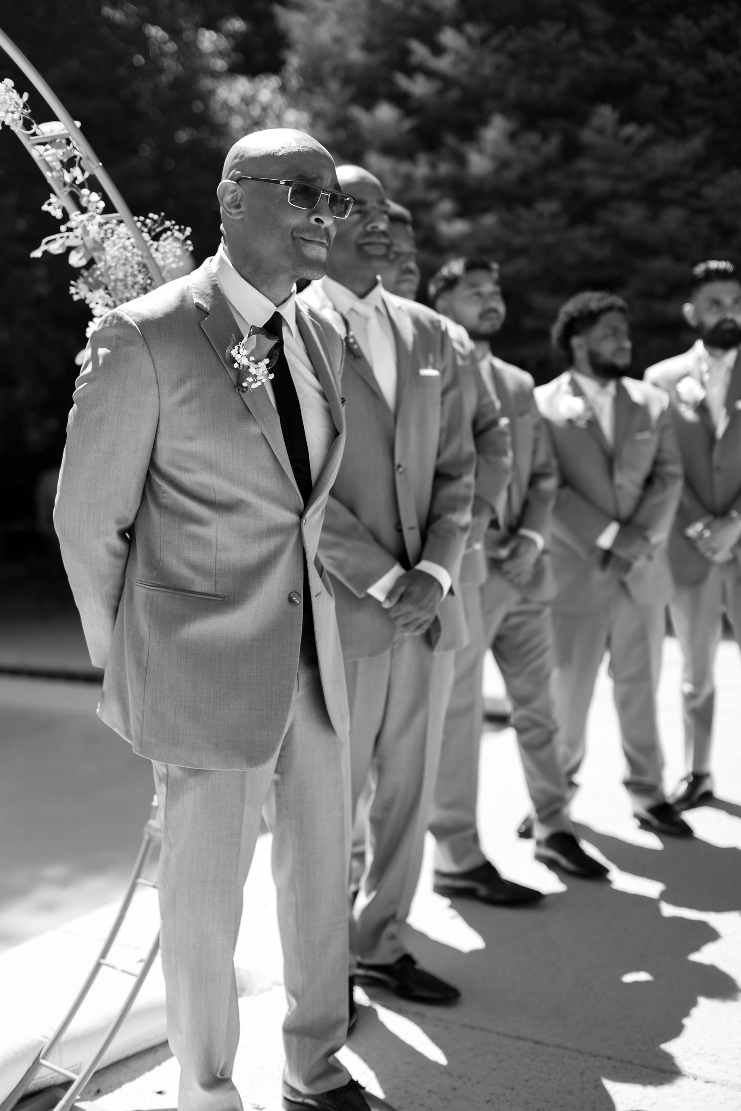 Groom and groomsmen in grey suits lined up at outdoor wedding ceremony in black and white.