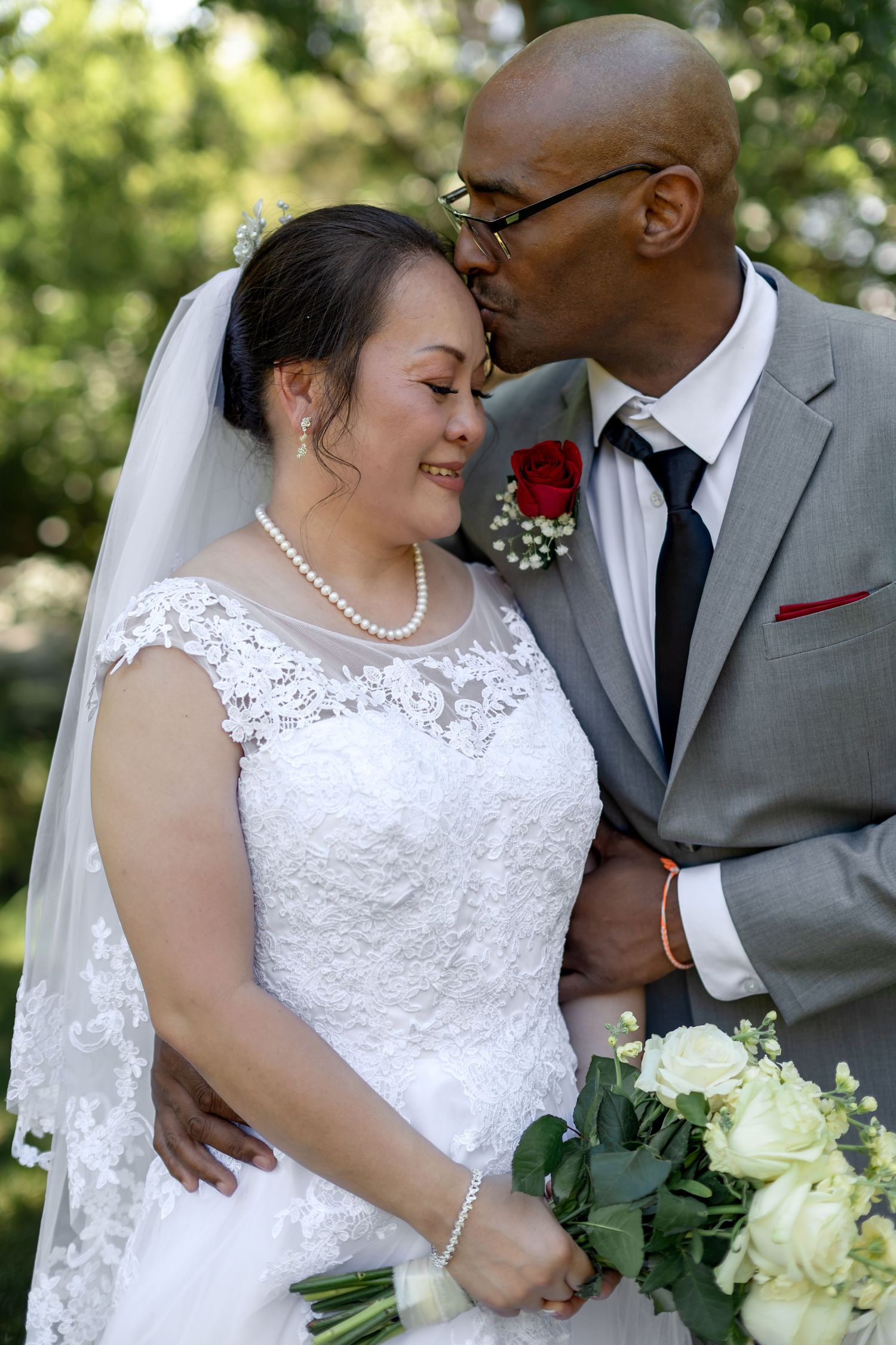 Groom kissing bride on the cheek as she smiles holding a white bouquet in a lace wedding dress.