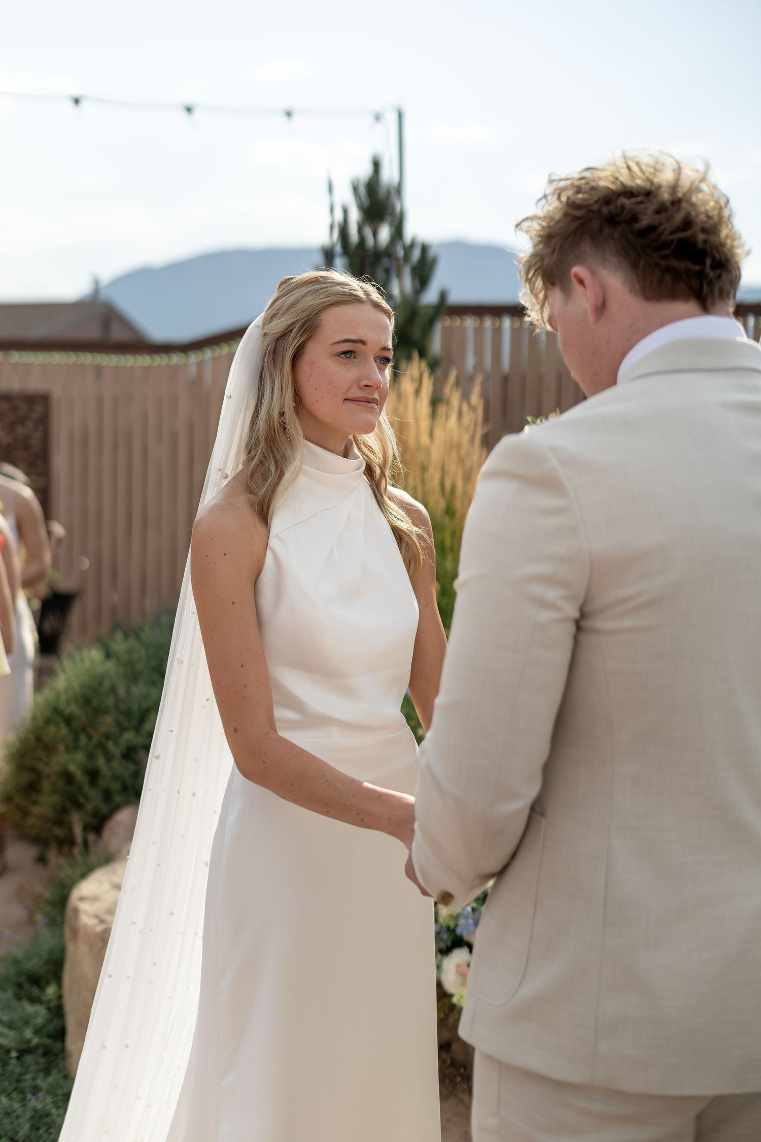 Blonde bride in elegant halter wedding gown holding hands with groom during outdoor wedding ceremony.