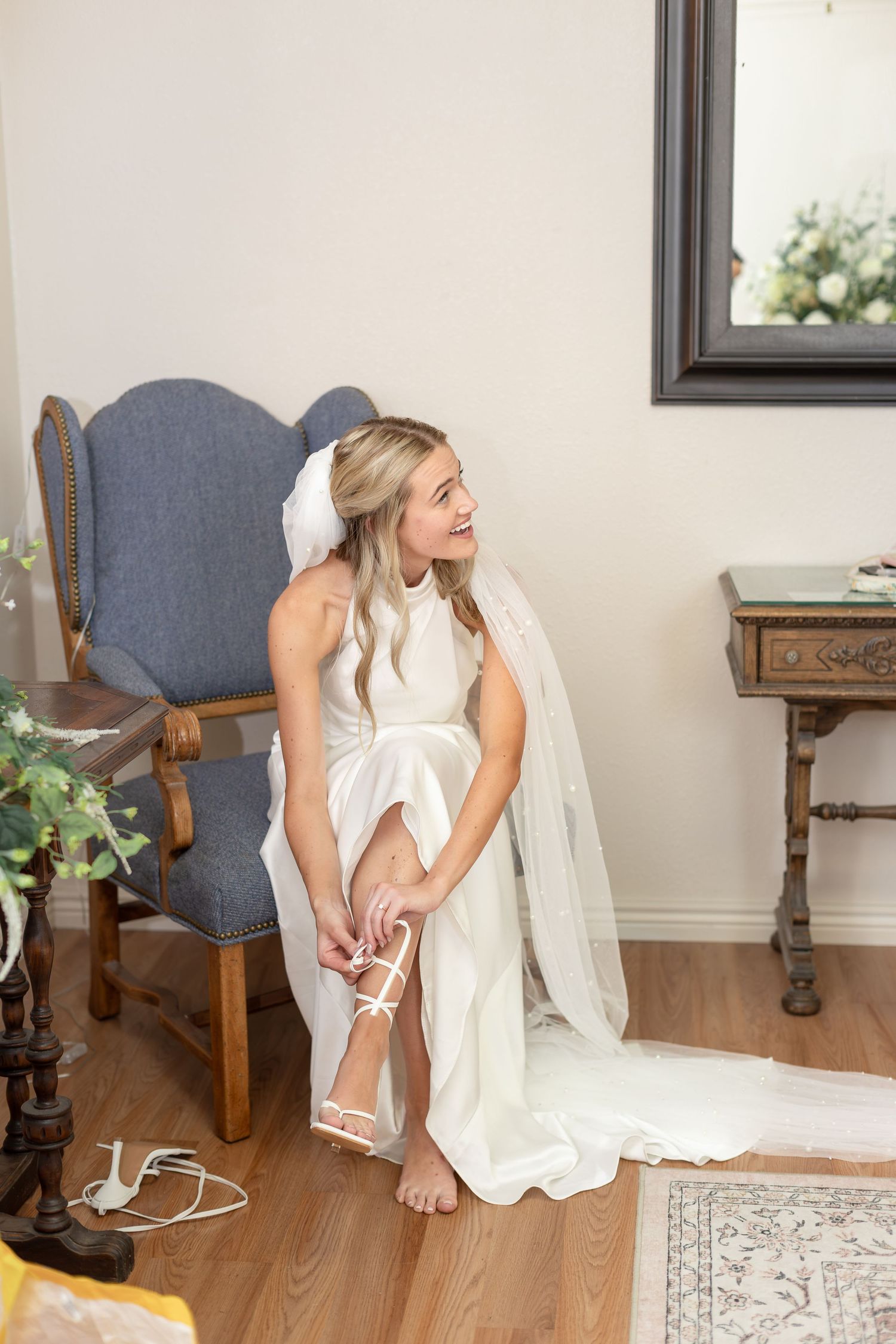 Smiling bride in white halter gown fastening strappy heels while seated in blue velvet chair.