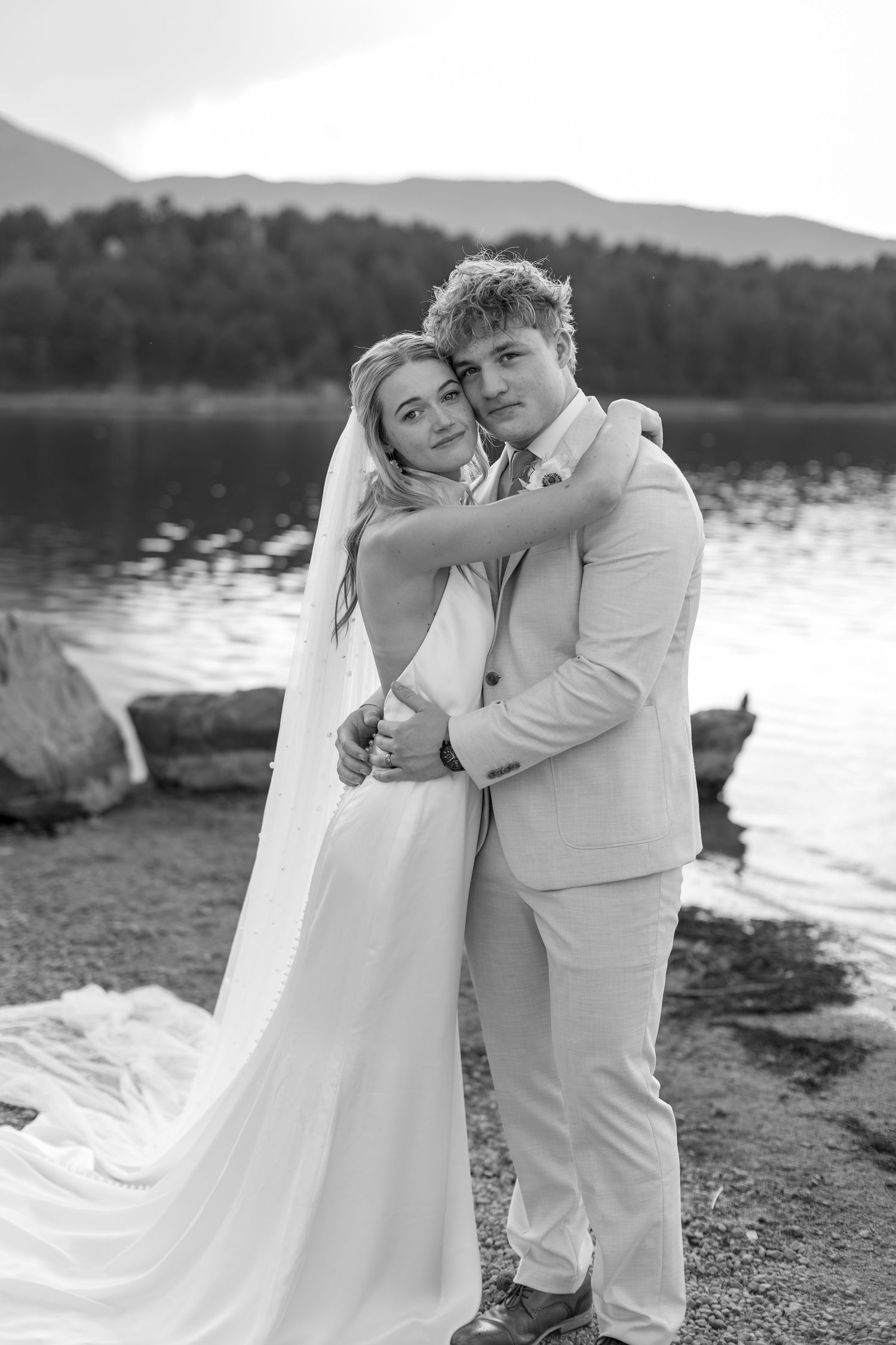 Black and white photo of a bride and groom embracing by a lake with mountains in the background.