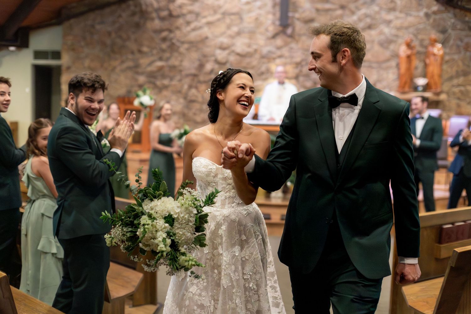 Bride and groom walk down church aisle smiling, holding hands, with guests applauding in a stone chapel.