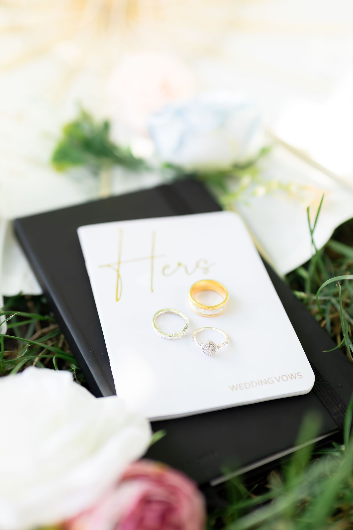 Gold wedding bands and diamond engagement ring rest on a white 'Hers' vow book surrounded by roses.