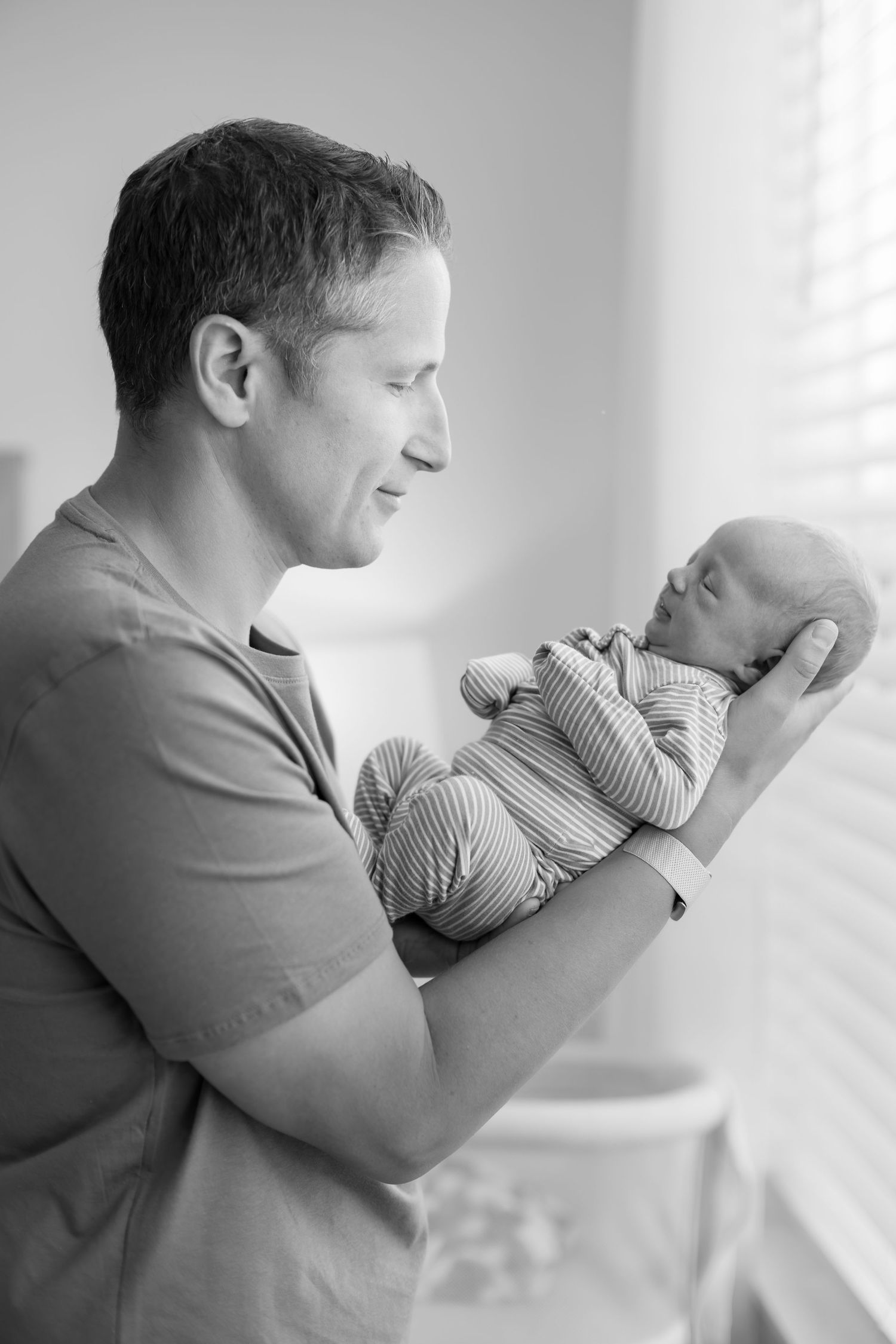 Black and white photo of smiling father tenderly holding newborn baby near bright window.