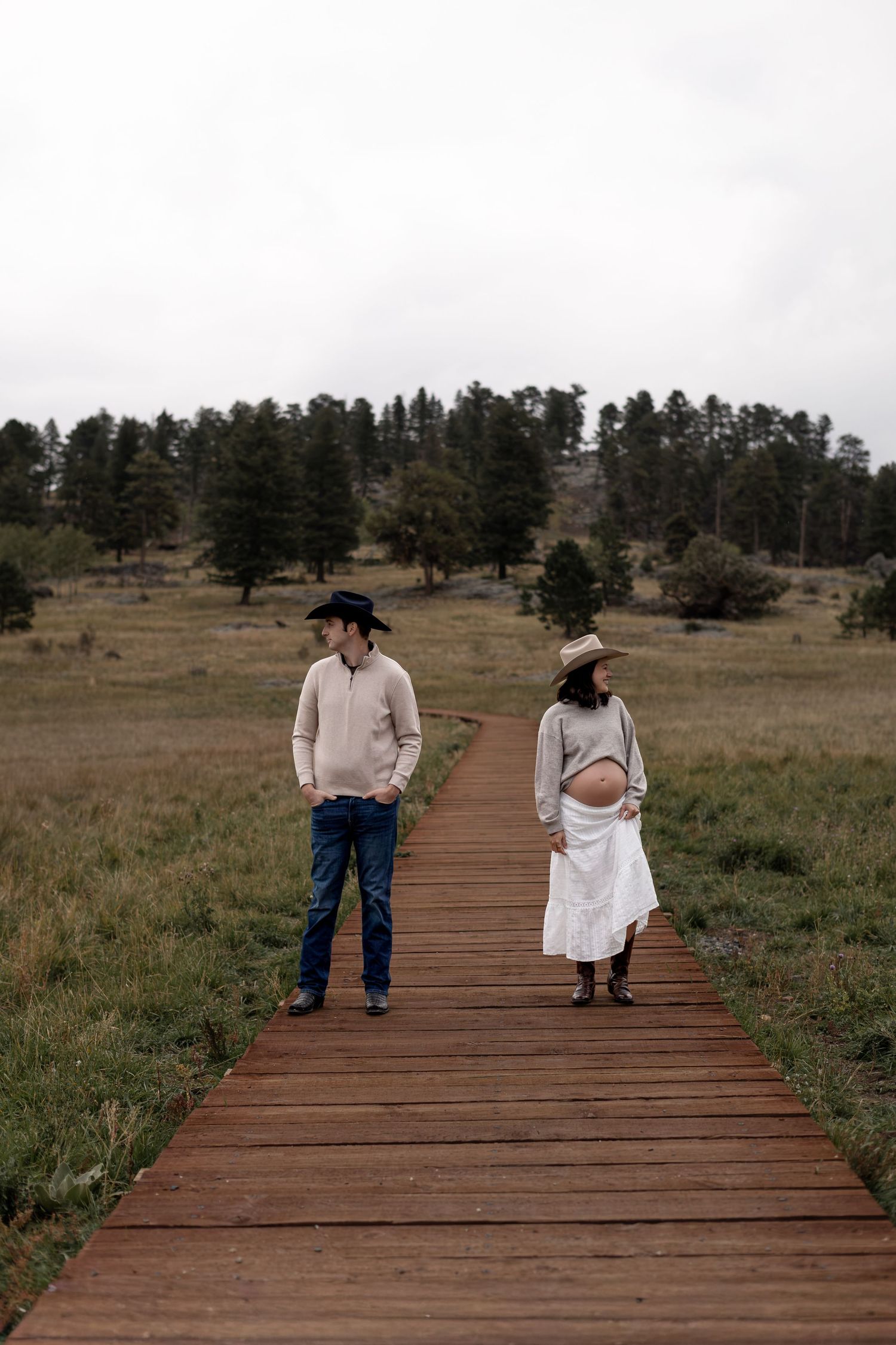 A couple stands apart on a wooden boardwalk path through a rural meadow, the woman visibly pregnant in a white skirt.