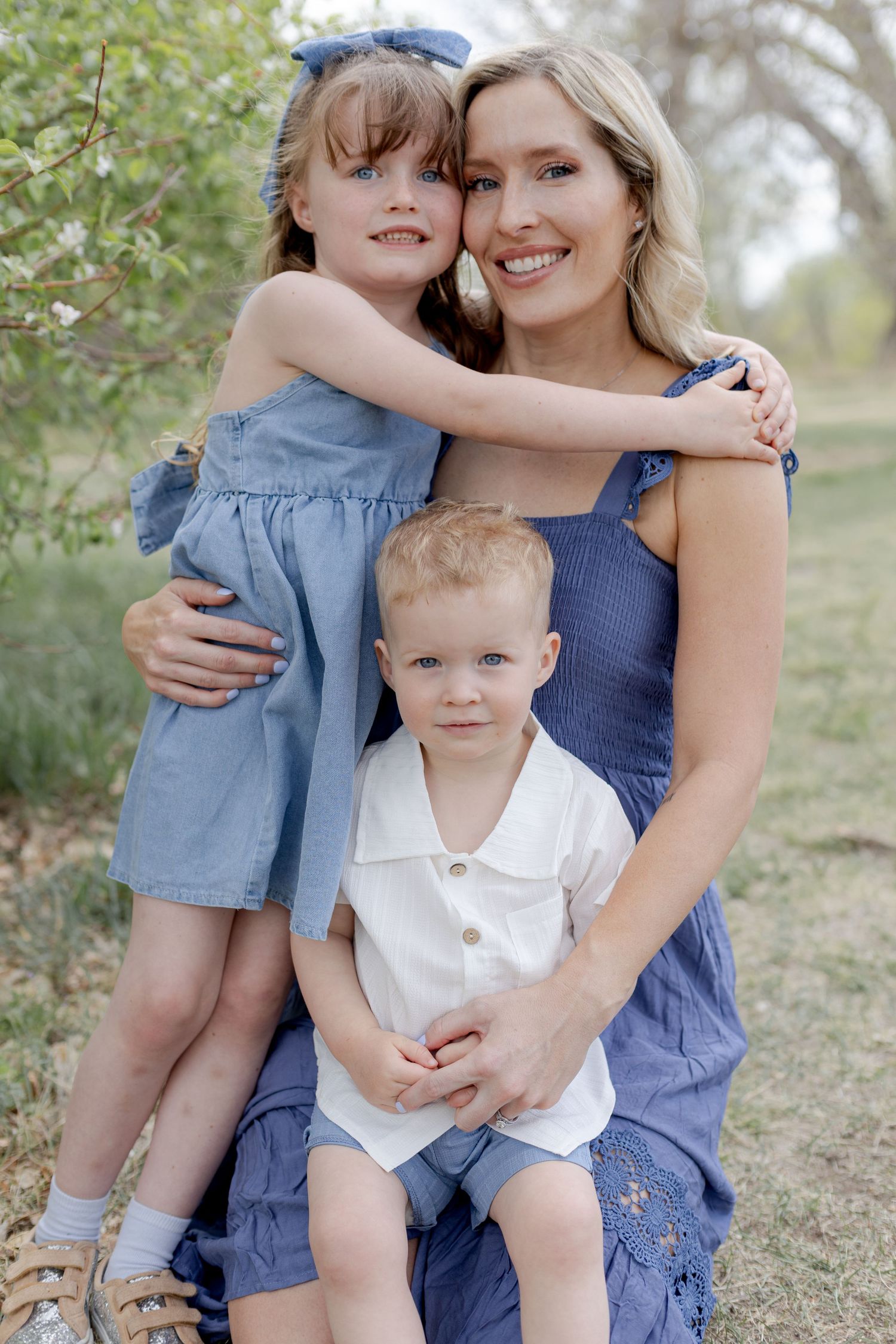 A smiling blonde woman in a blue dress poses outdoors with two young children hugging her.