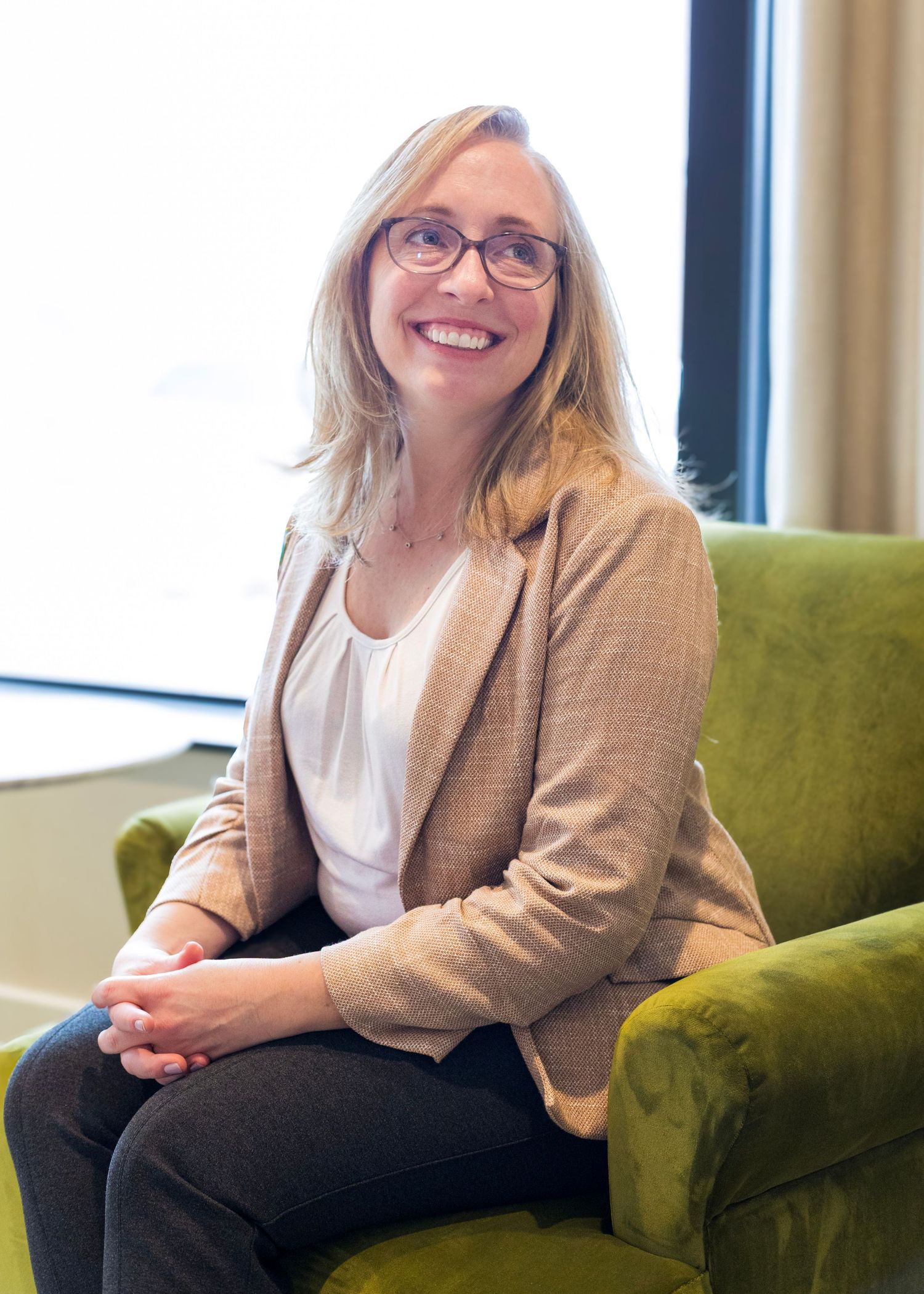 Professional wearing glasses and beige blazer sits comfortably on green velvet couch in office setting.