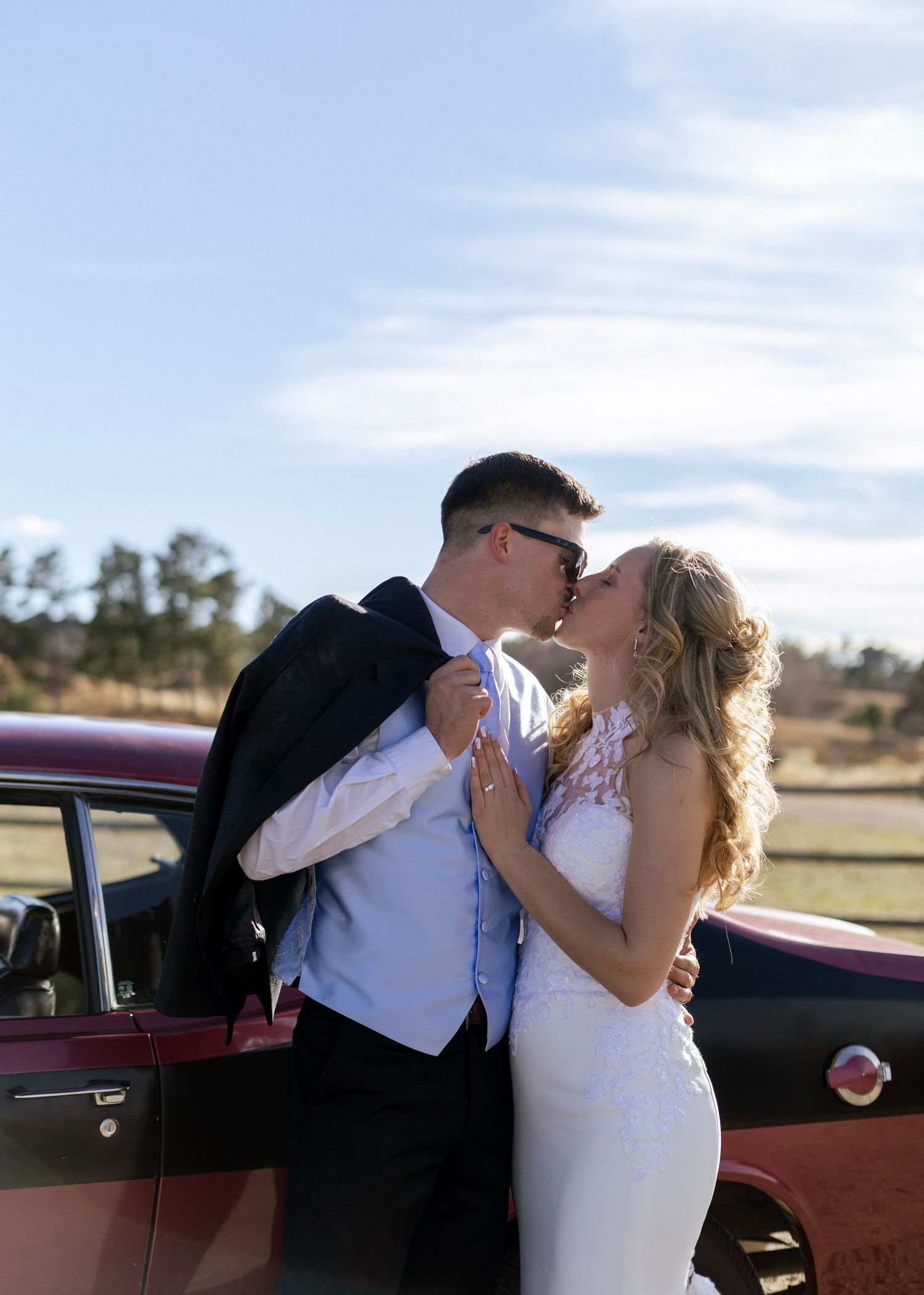 Bride and groom kissing beside a vintage red car under a blue sky at their outdoor wedding celebration.