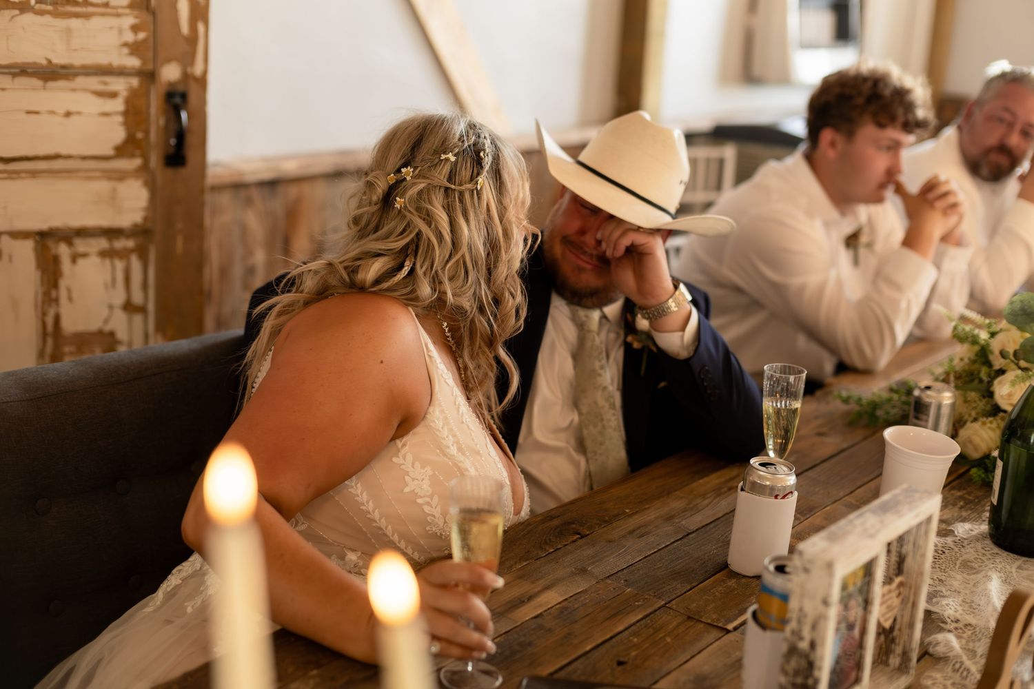 Bride and groom in cowboy hat share tender moment at candlelit rustic wooden wedding reception table.