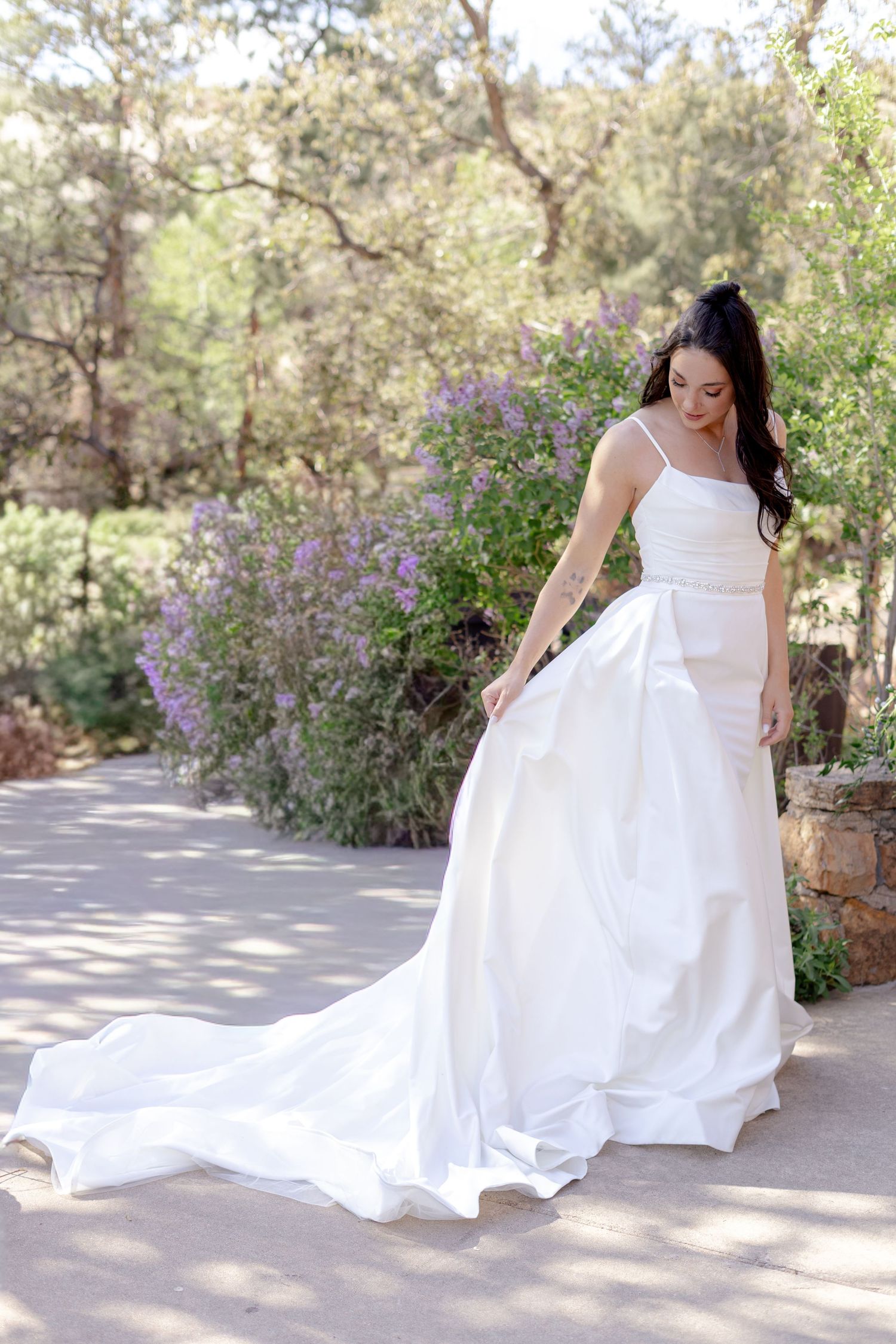 Elegant bride in a flowing white satin gown with train poses outdoors among purple blooming flowers and trees.