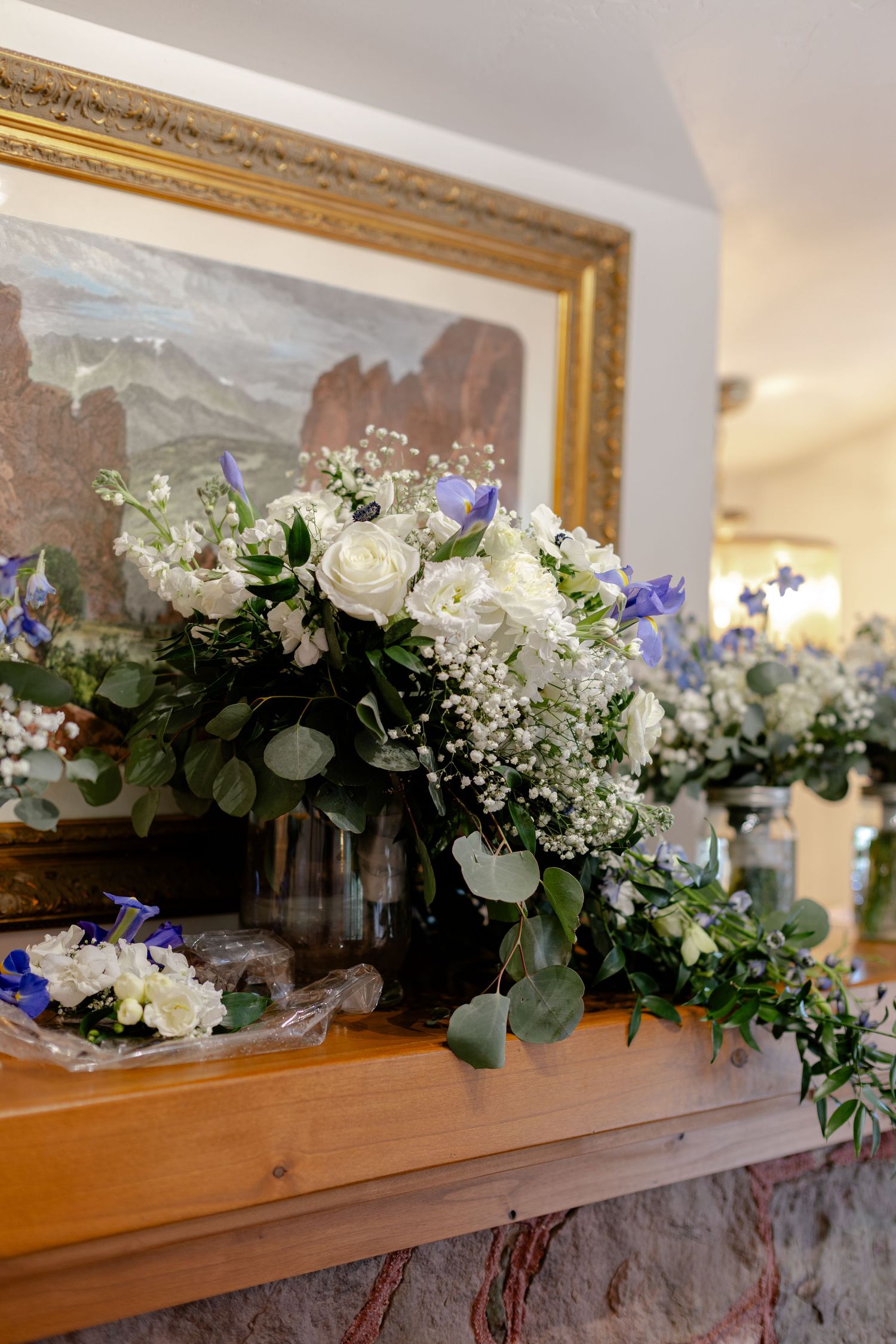Elegant wedding floral arrangement with white roses, blue flowers, and eucalyptus on a wooden mantle before a gold-framed painting.