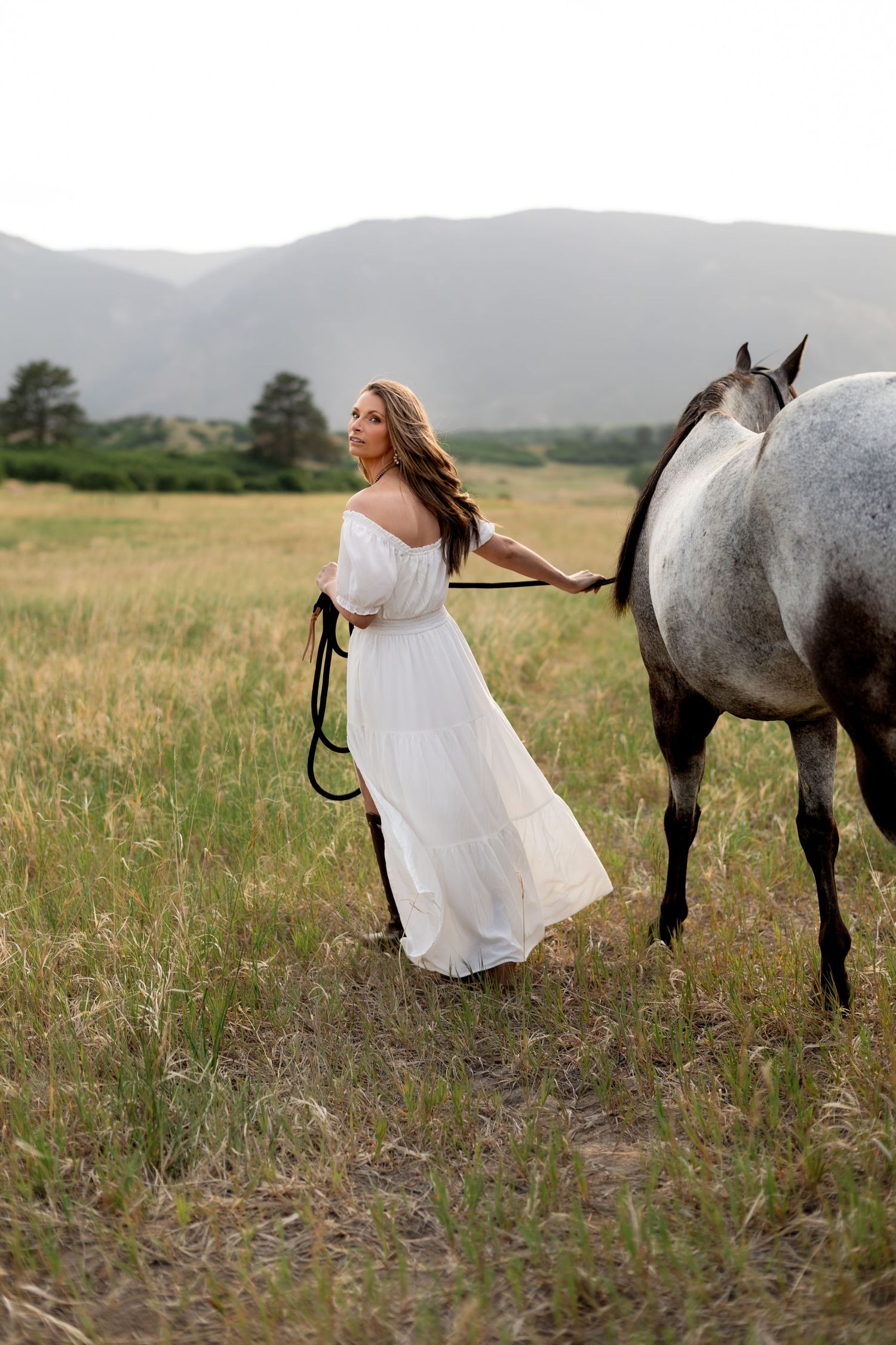 Woman in white dress leading a gray horse through a scenic mountain meadow at golden hour.