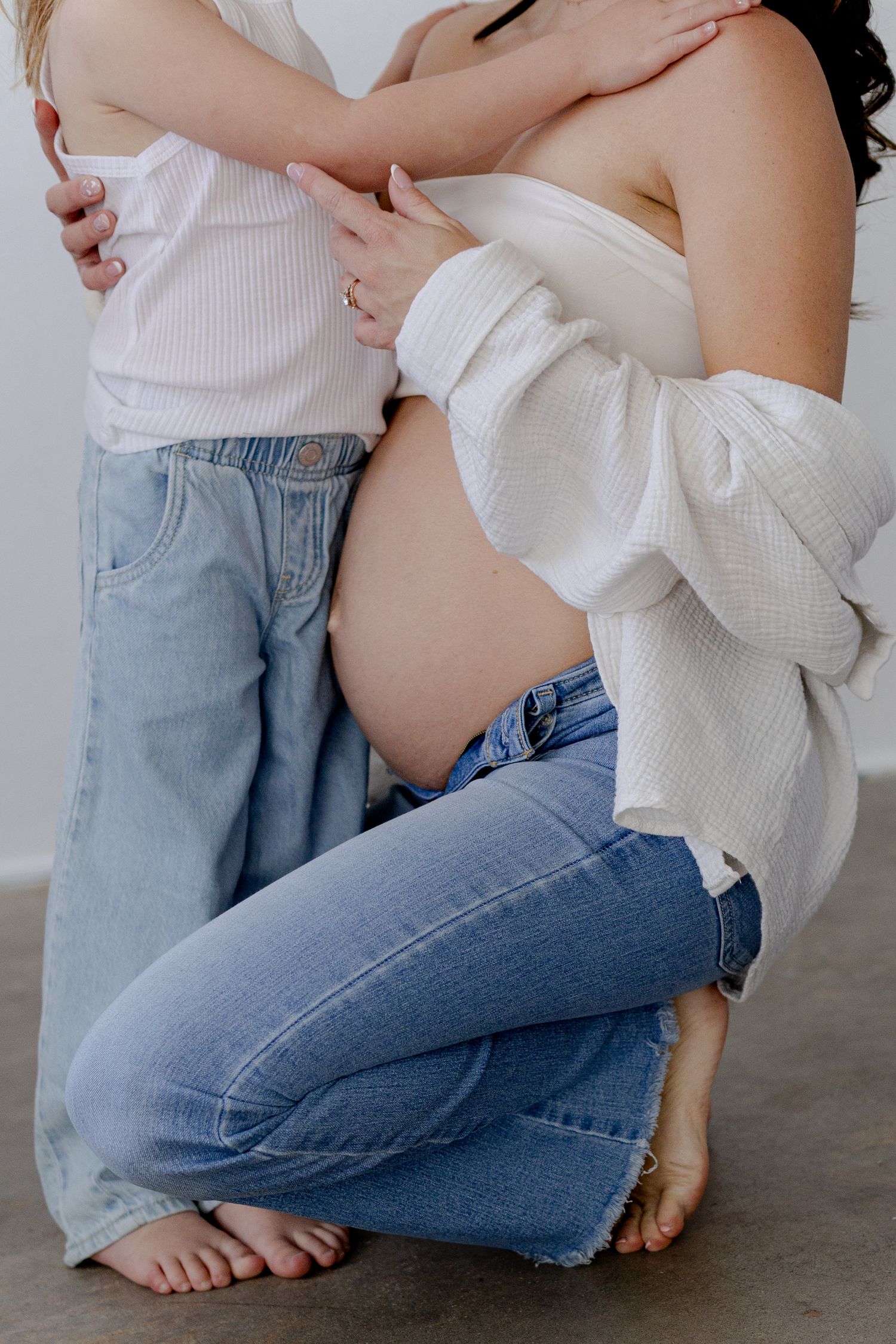 Pregnant woman in jeans kneeling while young child embraces her bare baby bump in a soft white studio setting.
