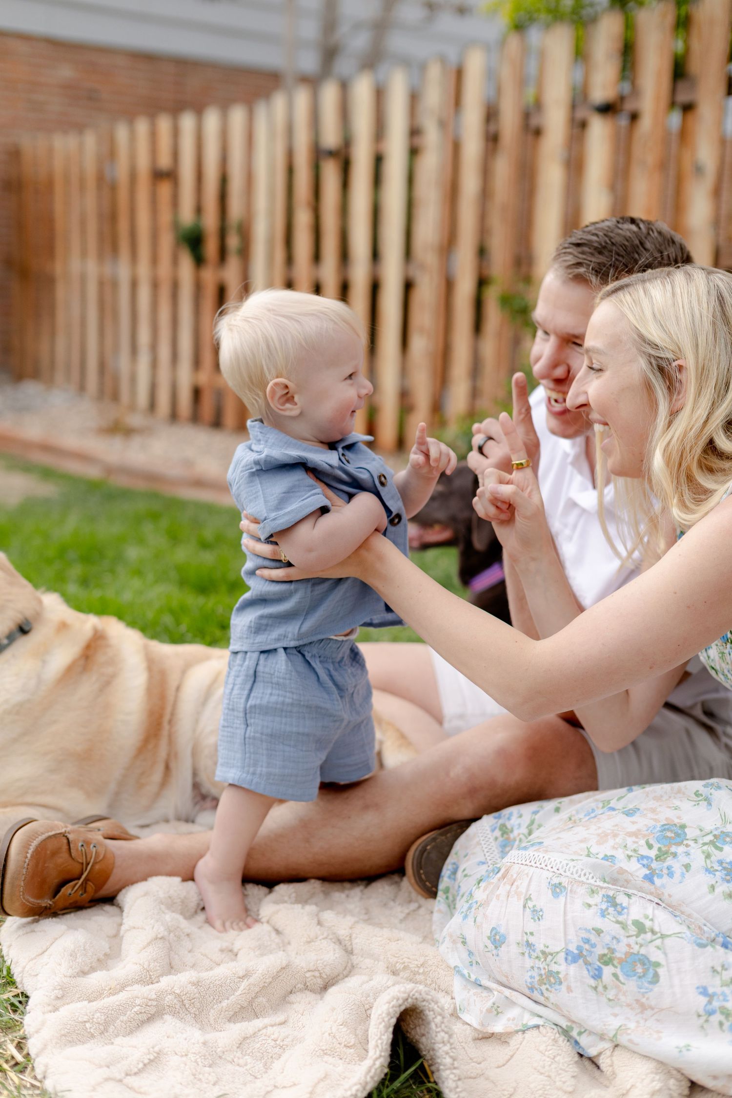 A happy family with a baby and golden retriever enjoying a sunny day on a blanket in their backyard.