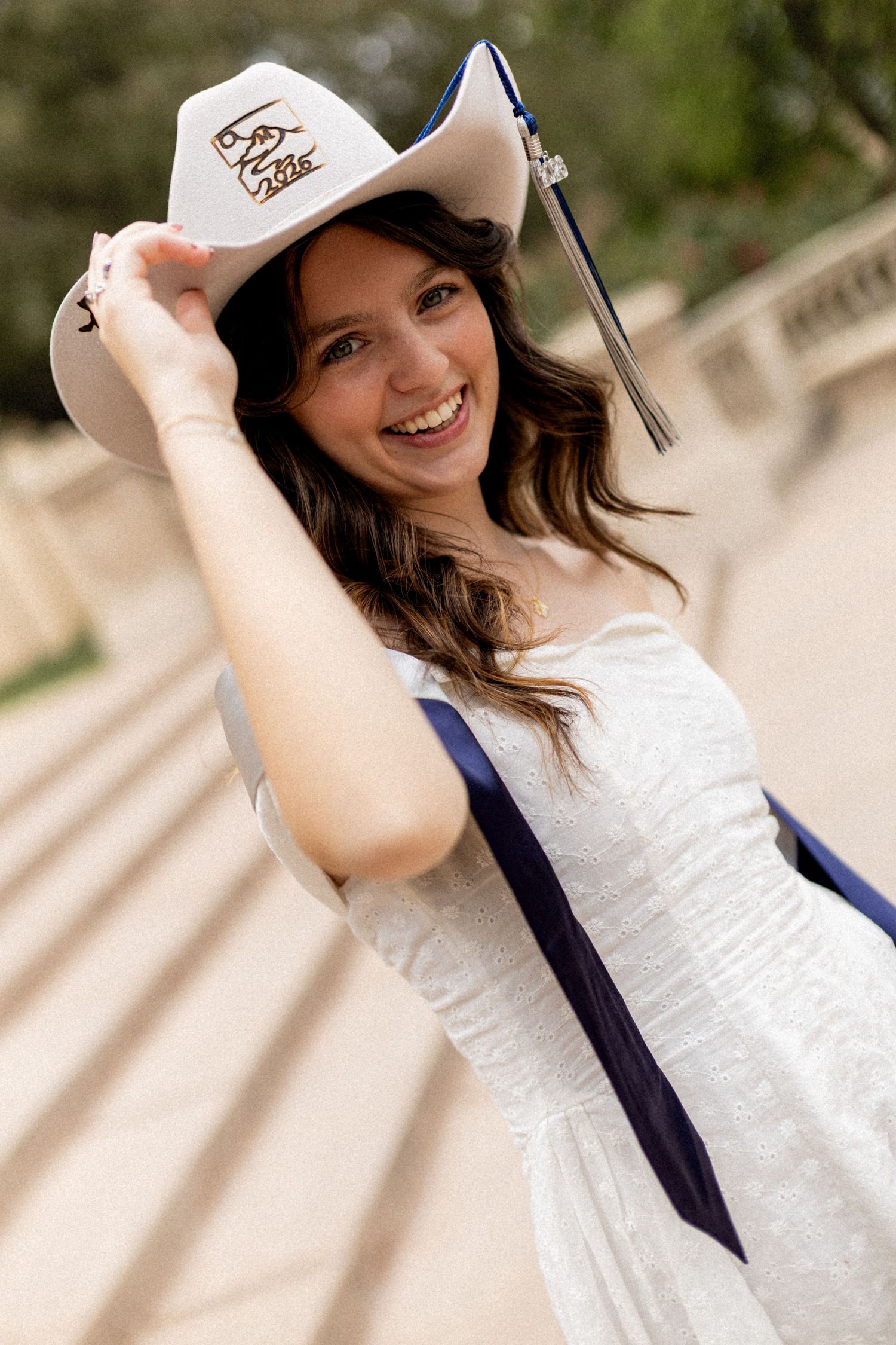 Happy graduate smiling in white dress wearing cowboy hat with blue graduation sash outdoors.
