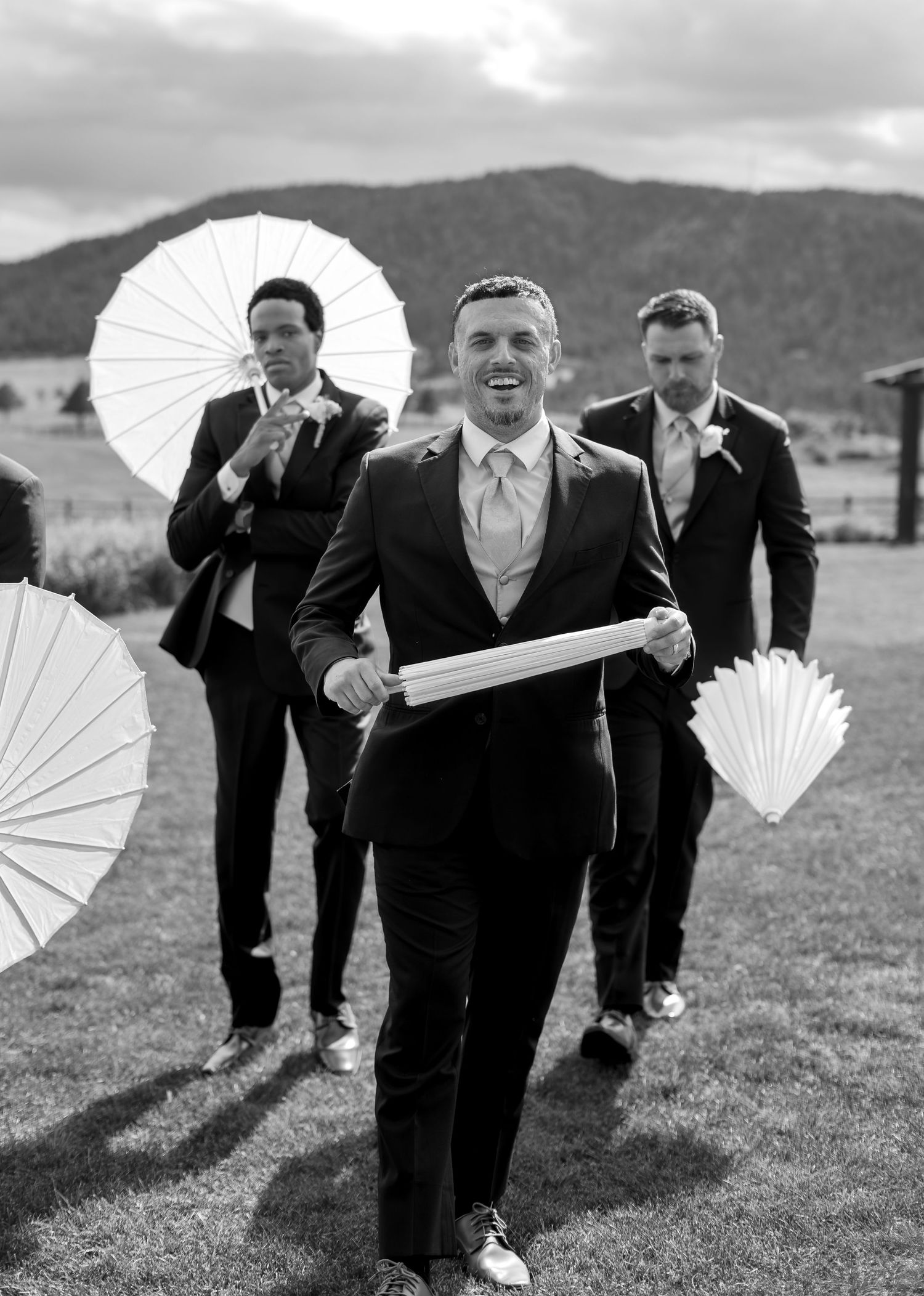 Smiling groom in black suit holding white fan leads groomsmen across field at wedding.