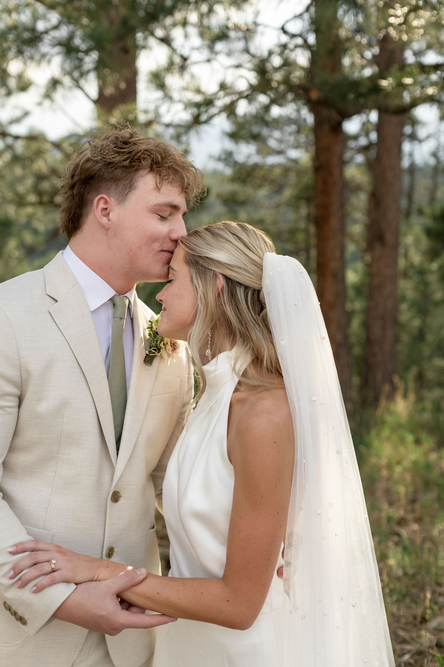 Groom in tan suit kisses bride on forehead as she holds his hands in a pine forest setting.