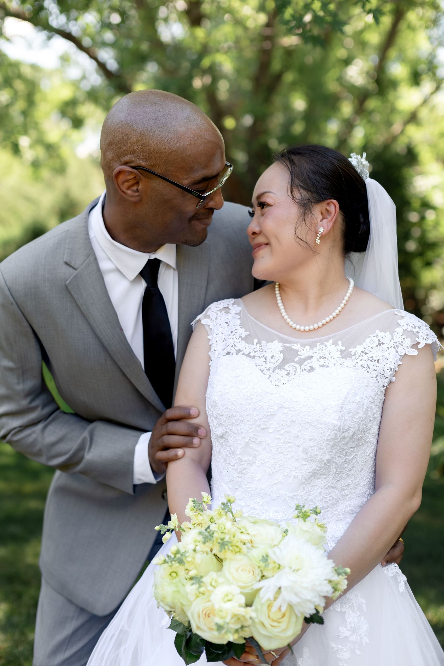 Interracial couple in wedding attire share a romantic moment outdoors, bride holding white rose bouquet.