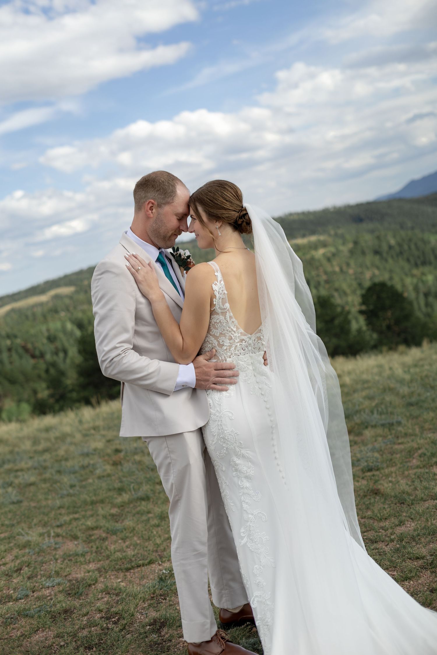 Bride and groom embrace forehead-to-forehead in a field with mountain views on their wedding day.