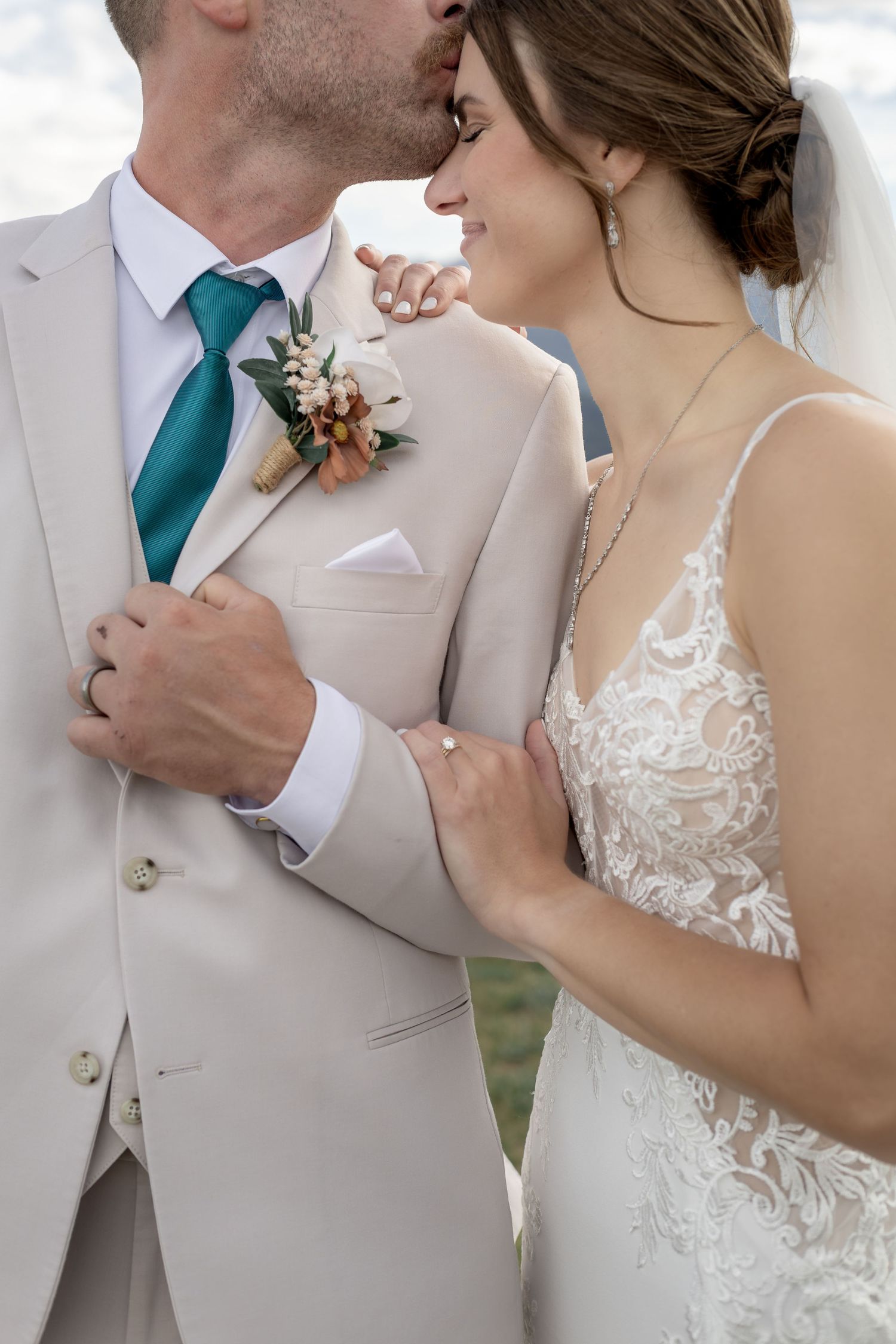 Groom in beige suit with teal tie kisses bride in lace gown as she smiles and holds his lapel.