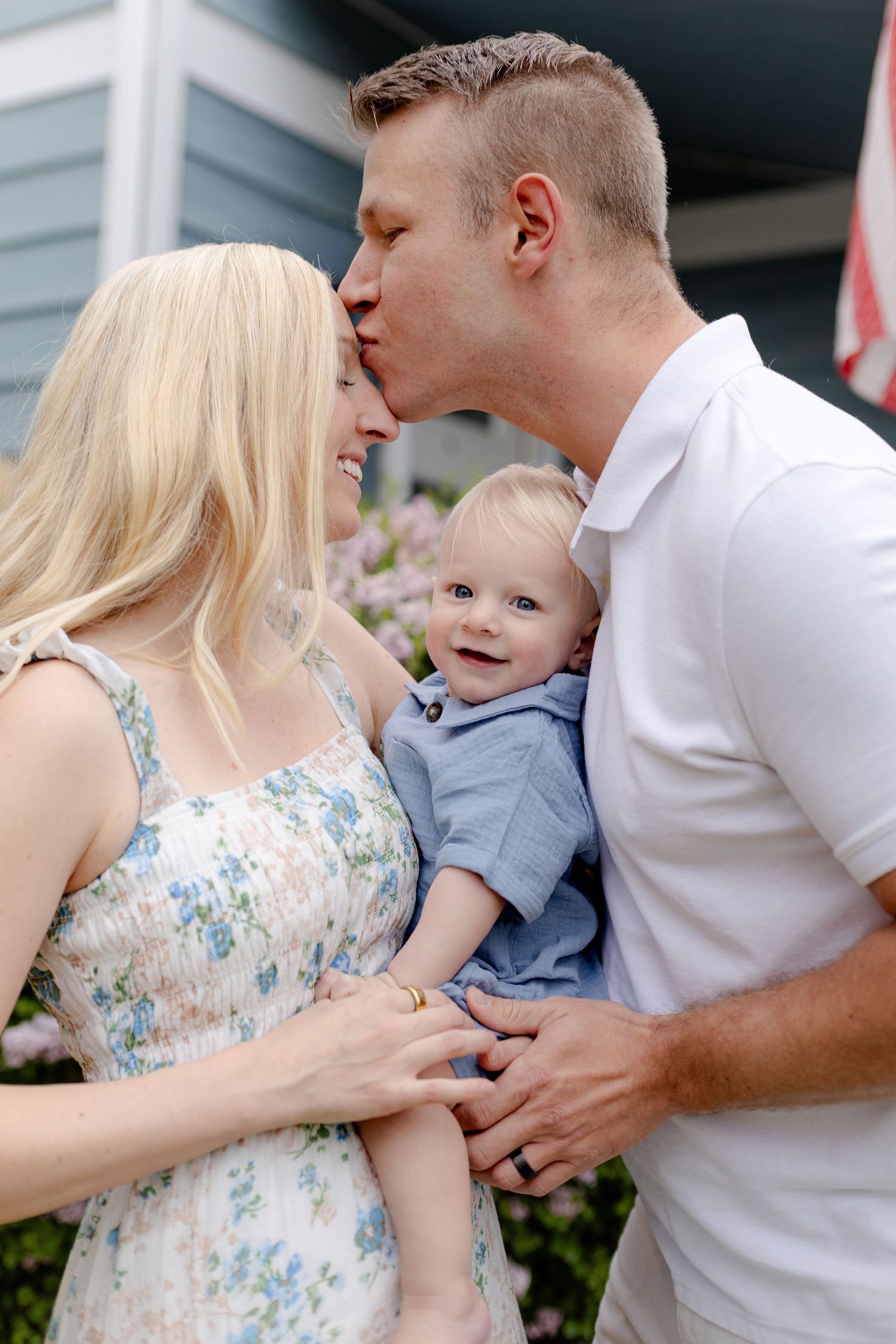 A couple shares a tender kiss over their smiling baby boy outdoors near a house with flowers.