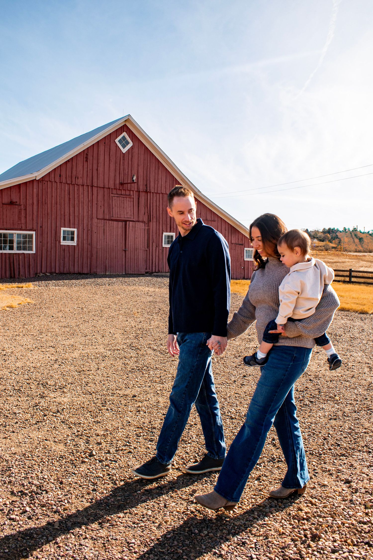 A young family walks hand-in-hand near a red barn on a sunny day, with the mother carrying a toddler.