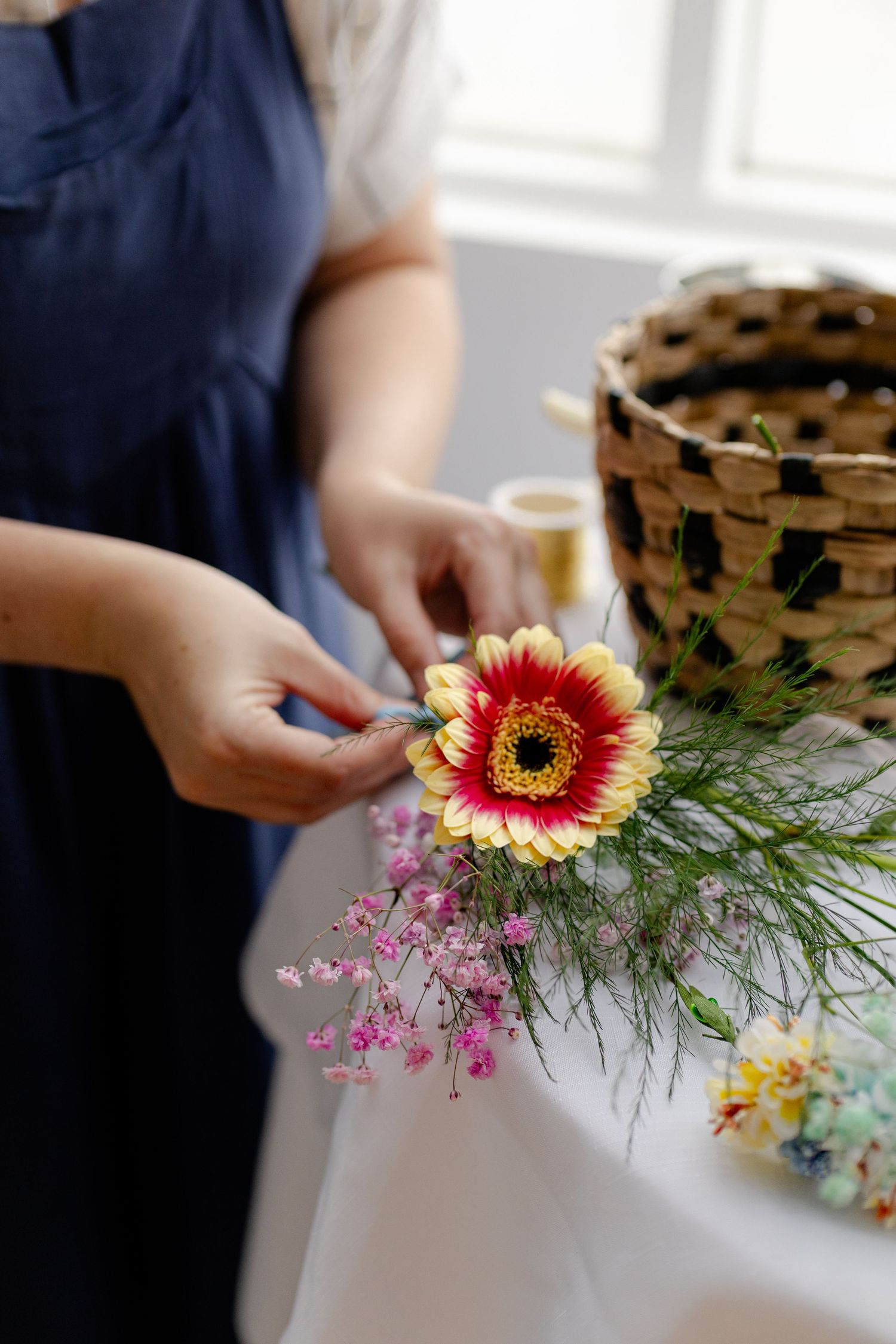 Woman in blue apron arranging a red and yellow gerbera daisy bouquet with pink flowers on a white table.