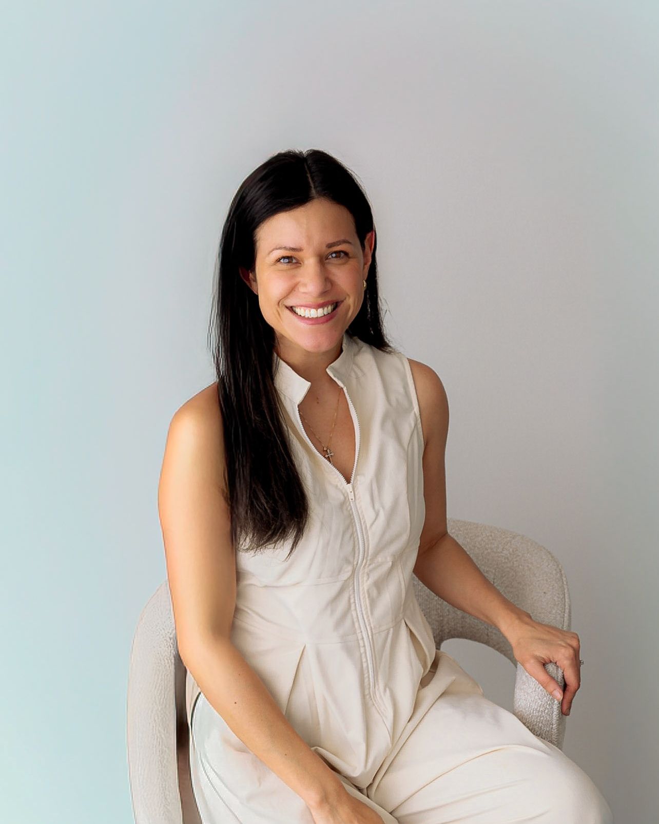 Smiling woman with dark hair wearing a white sleeveless outfit seated in a modern chair against a light blue background.