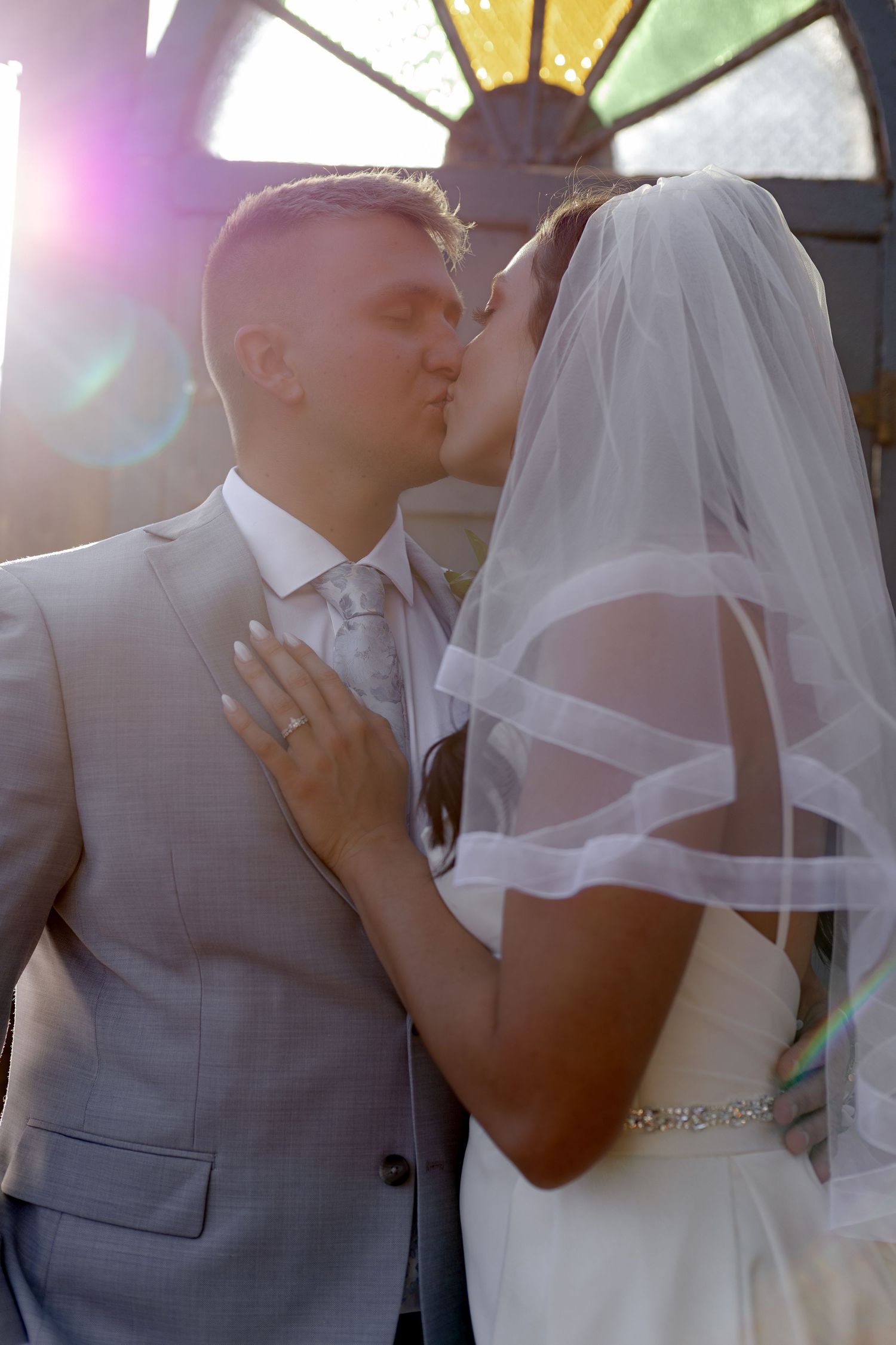 Newlyweds share a first kiss at the altar with stunning stained glass window and golden sunlight lens flare.