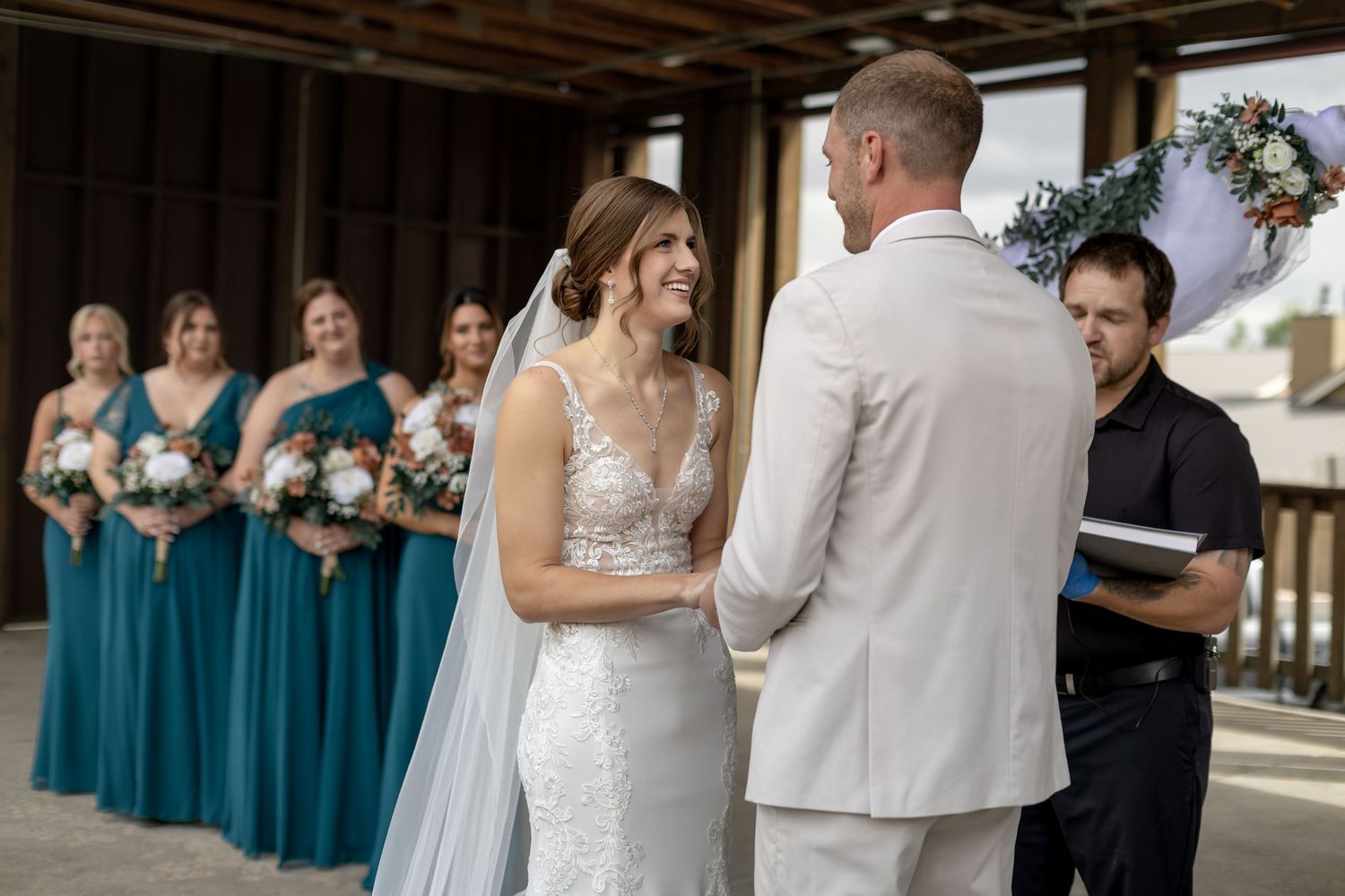 Bride smiling at groom during outdoor wedding ceremony with teal-dressed bridesmaids holding bouquets in background.