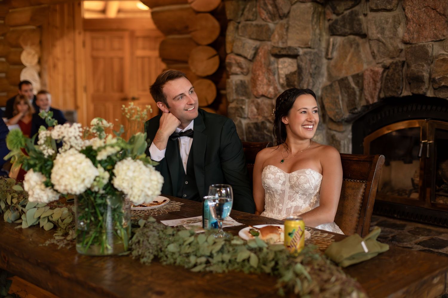 Bride and groom smiling at sweetheart table with floral centerpiece in rustic log cabin wedding venue.