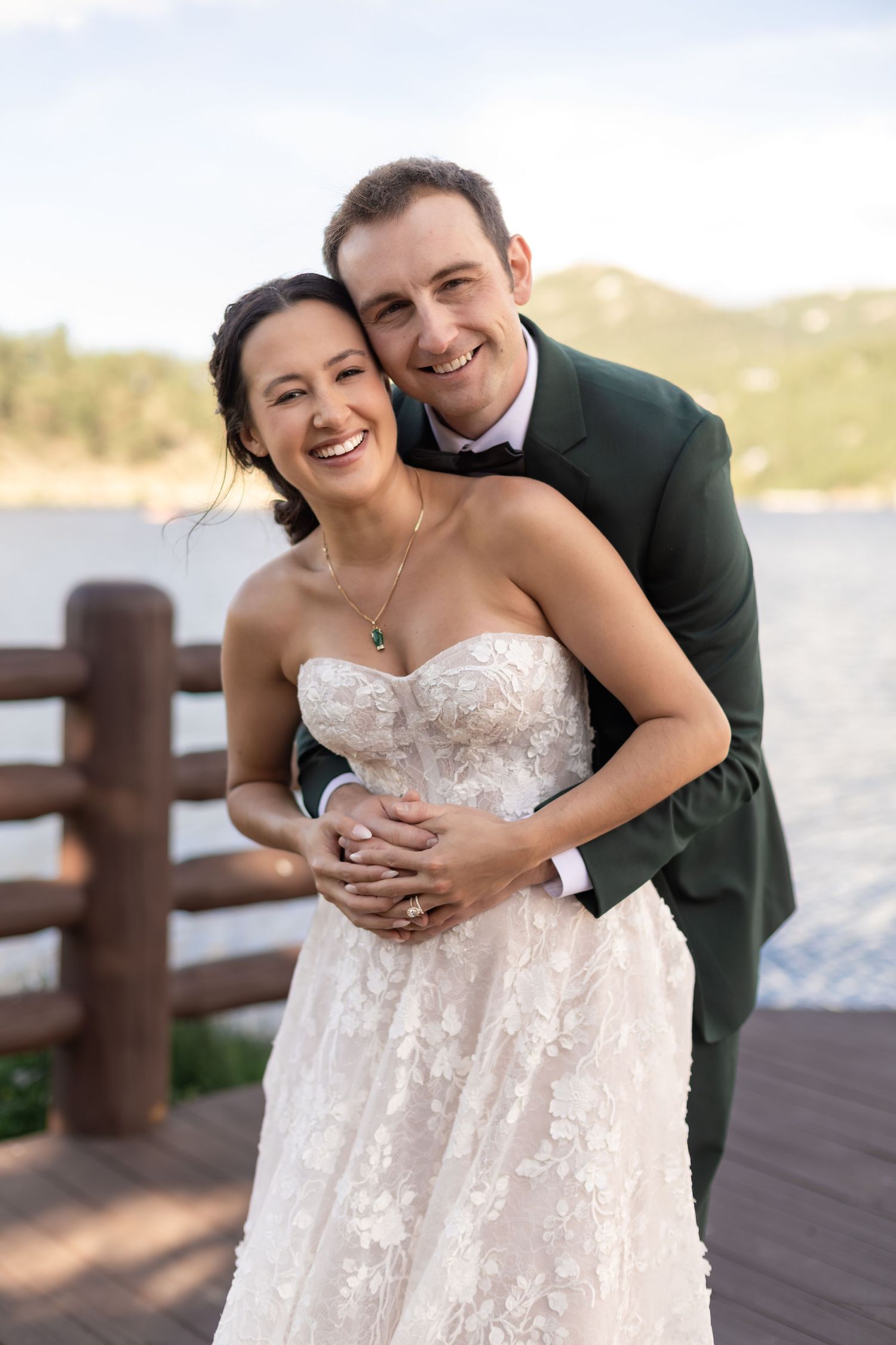 Smiling bride and groom embrace on a lakeside deck, she in a strapless lace gown, he in a dark green suit.