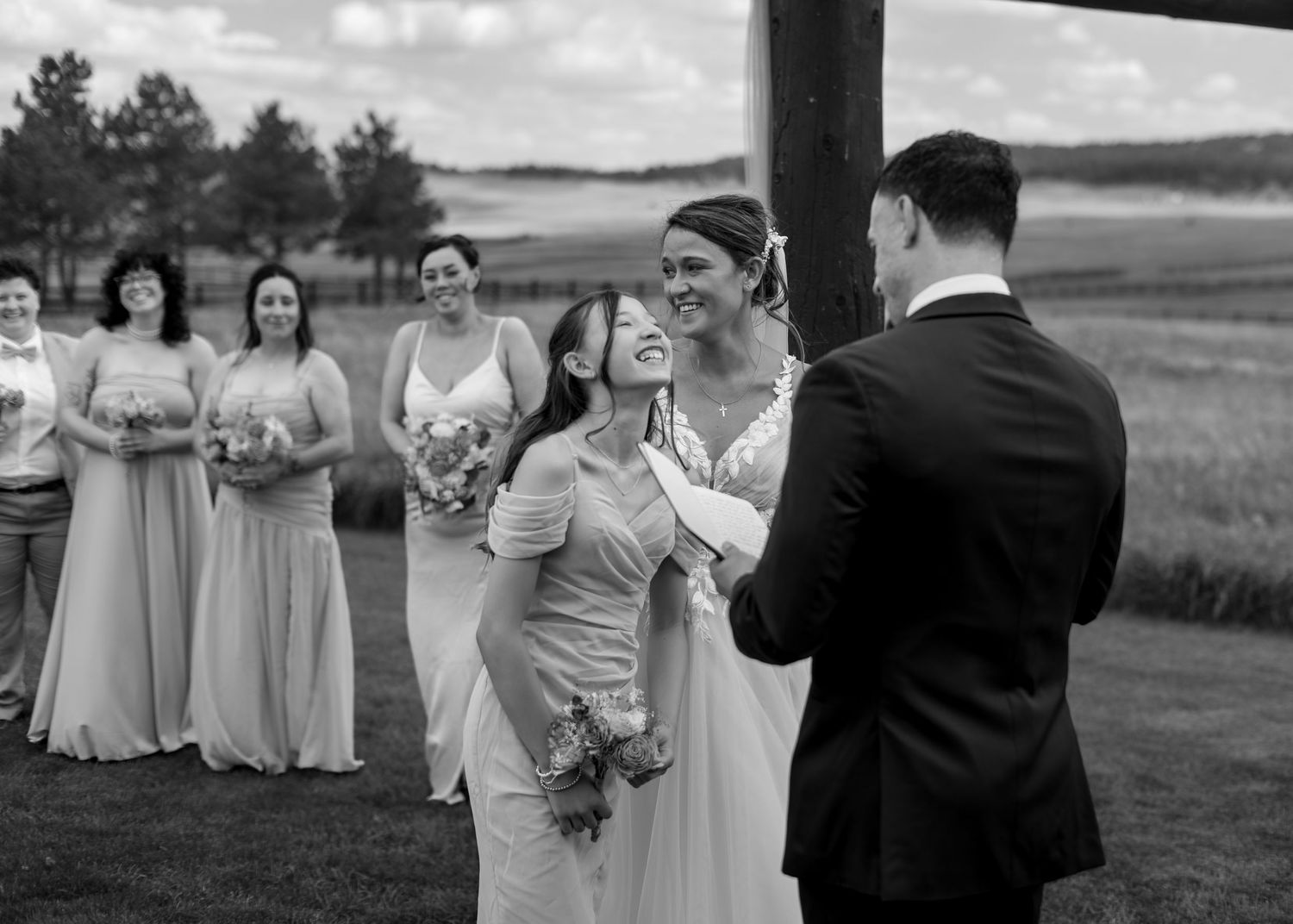 Black and white wedding ceremony photo of bride hugging a guest while groom watches, bridesmaids in background.