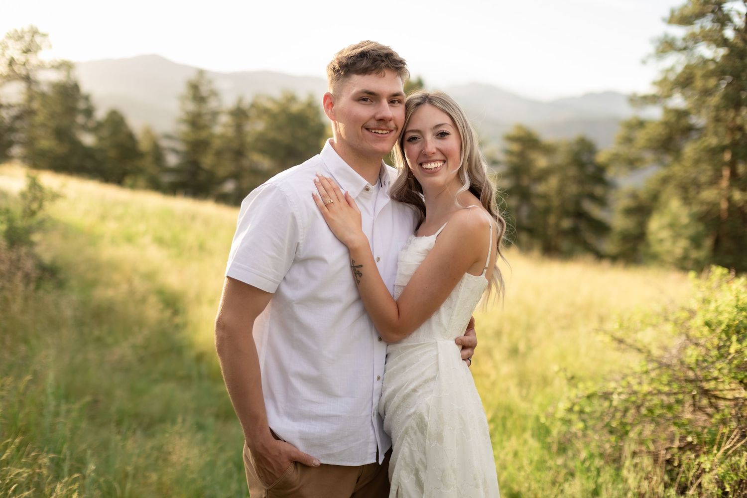 Couple smiling together in a sunny mountain meadow, man in white shirt and woman in white dress, with pine trees and hills.