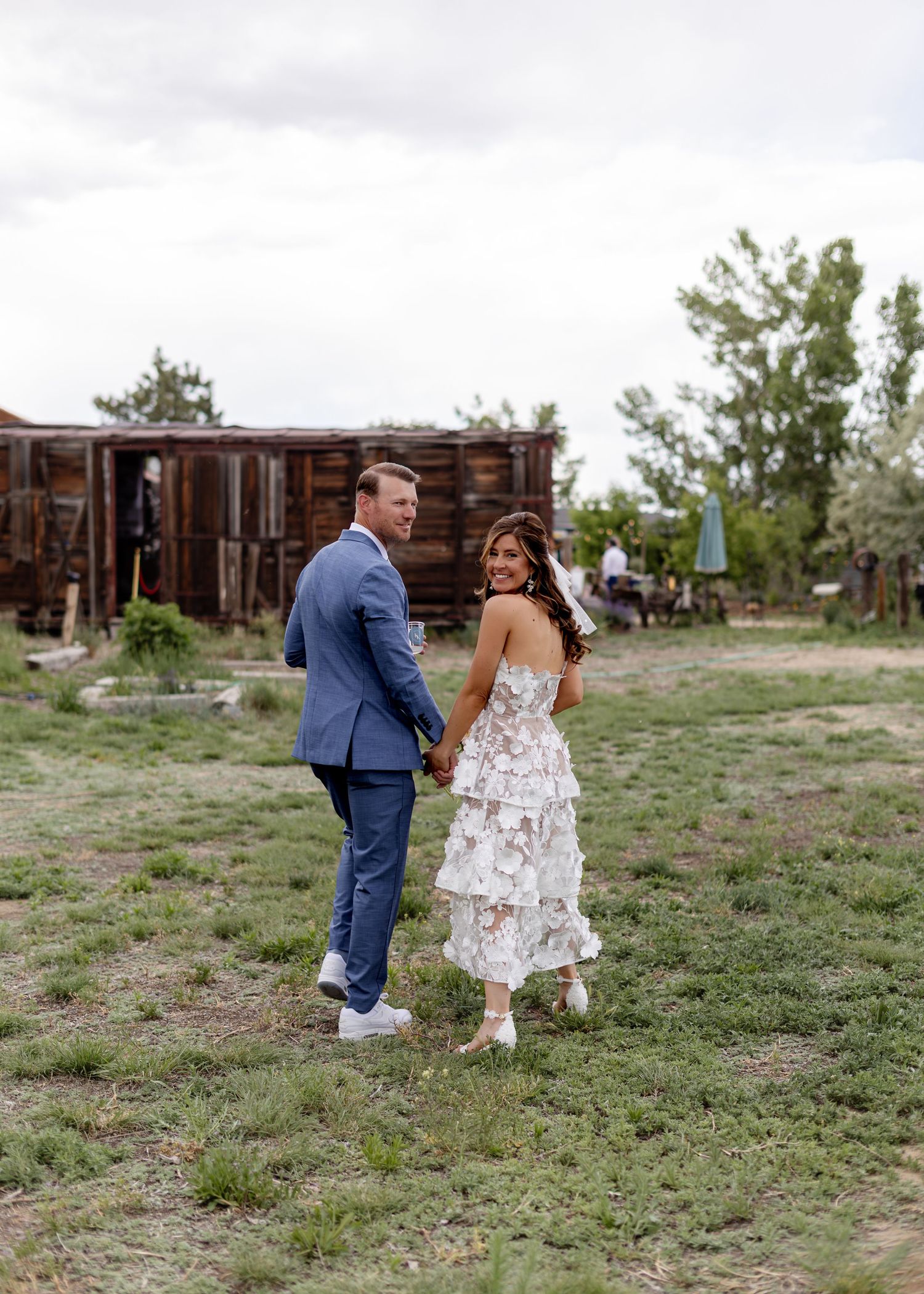 Couple holding hands walking outdoors, groom in blue suit, bride in white ruffled dress, rustic barn background.