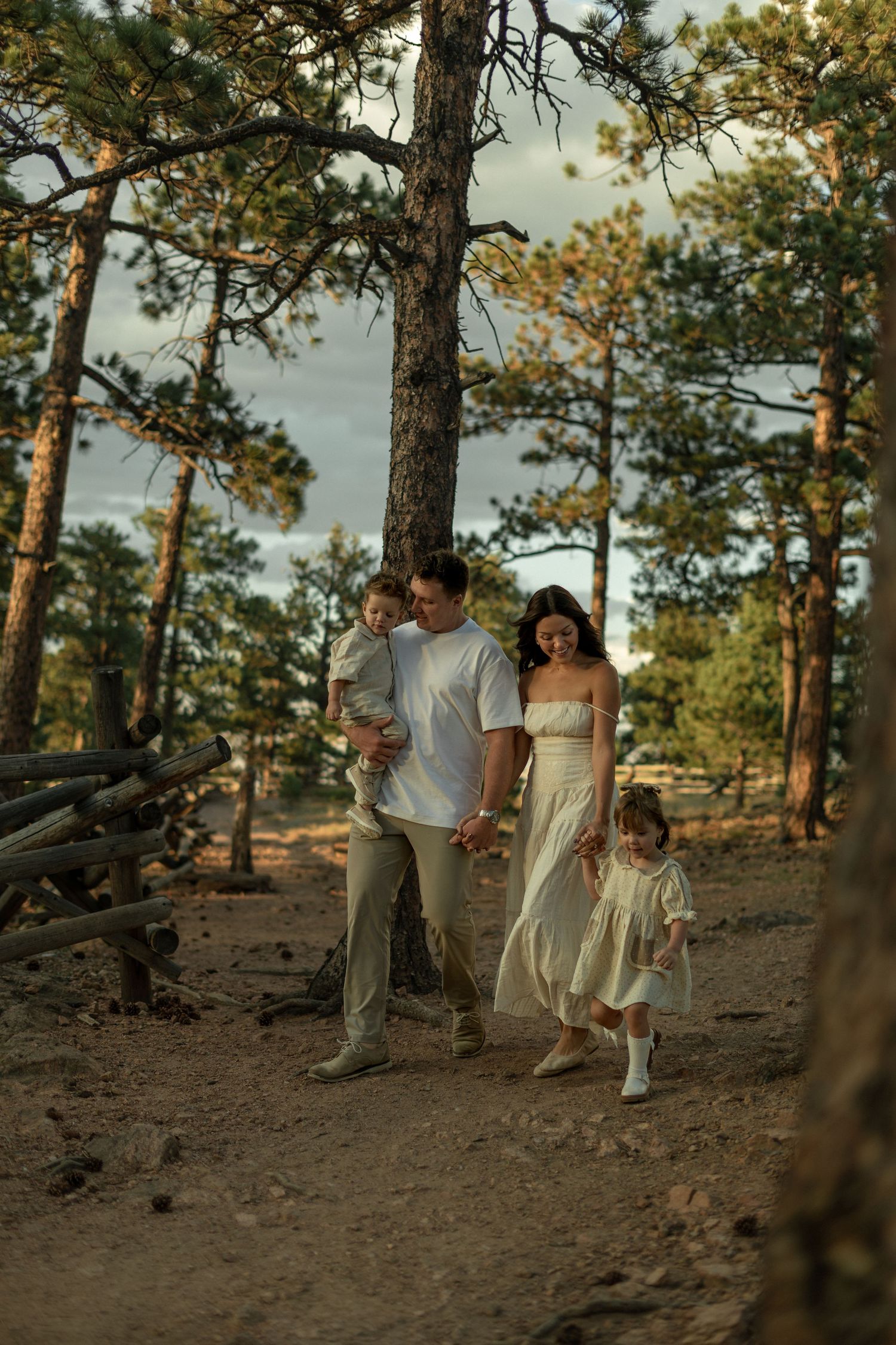 Family of four walking through a pine forest on a sandy path during golden hour, dressed in neutral tones.