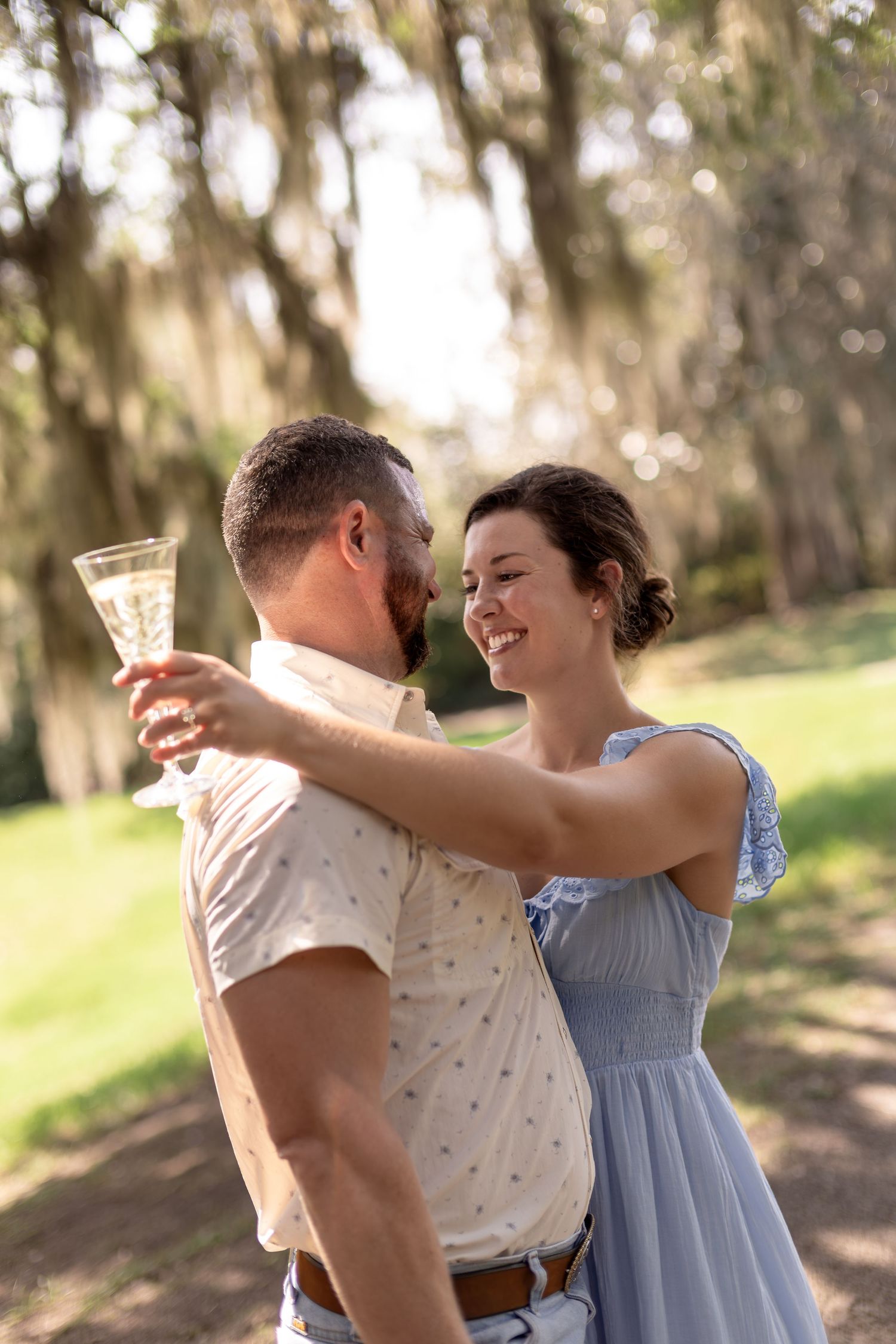 A smiling couple embraces and toasts with champagne glasses in a sunlit park with hanging moss trees.