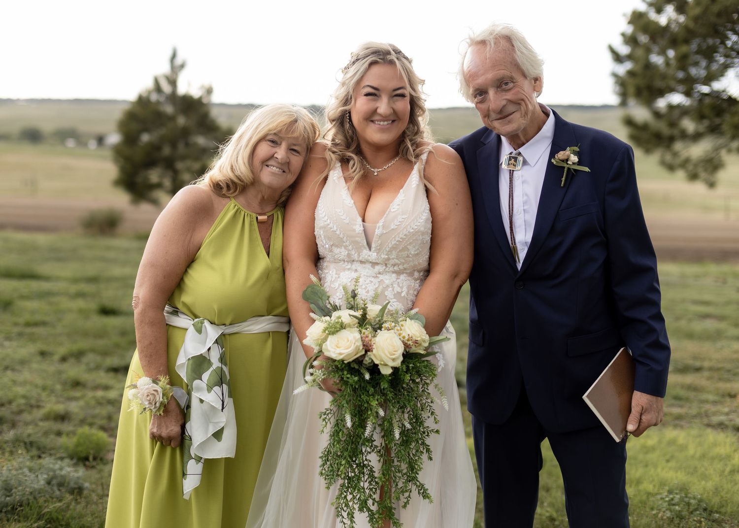 Bride with bouquet poses with parents in green and navy attire at outdoor wedding.