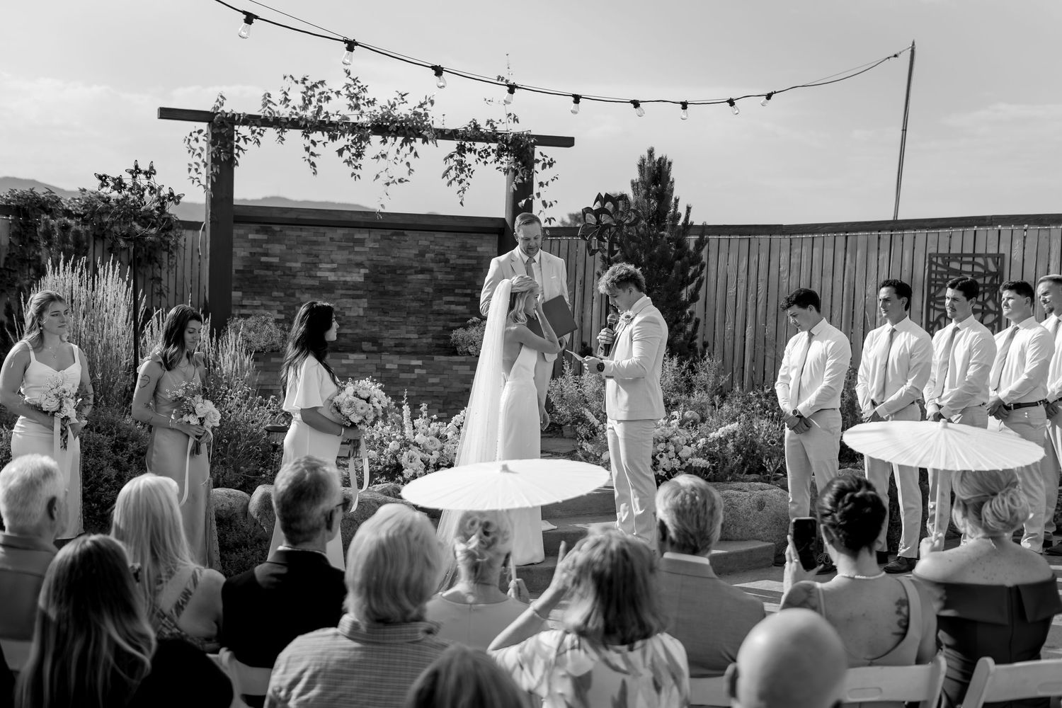 Black and white outdoor wedding ceremony with couple at altar, bridesmaids, groomsmen, and seated guests under string lights.