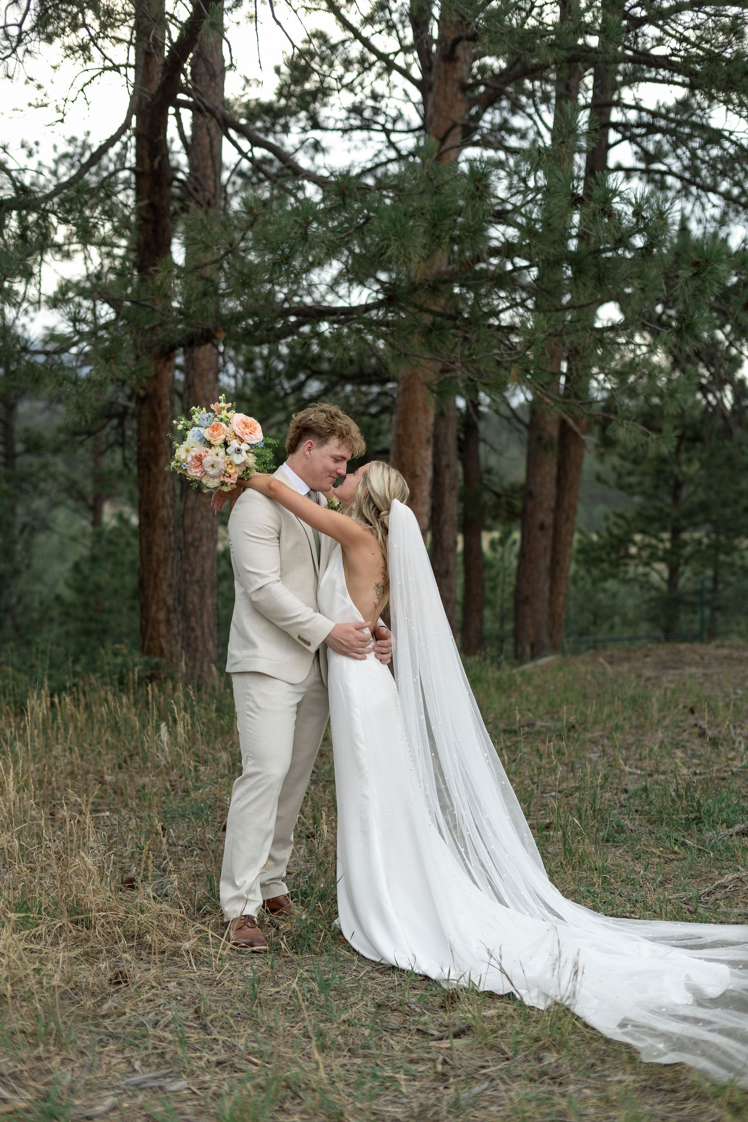 Bride and groom embrace under pine trees, bride in white gown with long veil holding colorful bouquet, groom in tan suit.